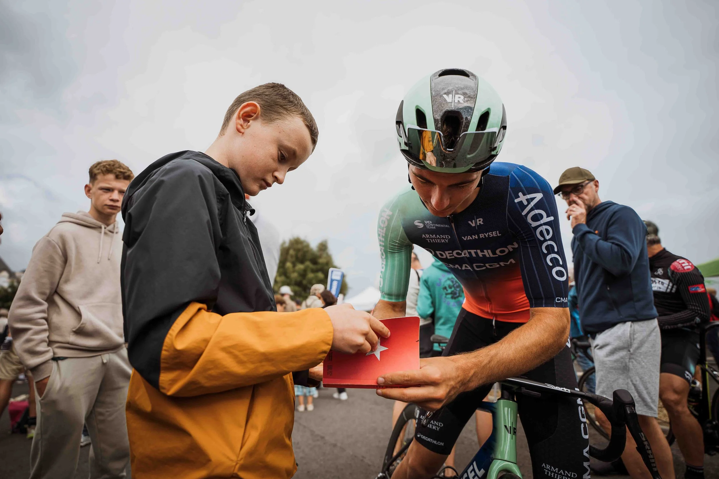 Un cycliste professionnel autographié par un jeune homme lors d'un événement public, entouré d'autres personnes sous un ciel nuageux.