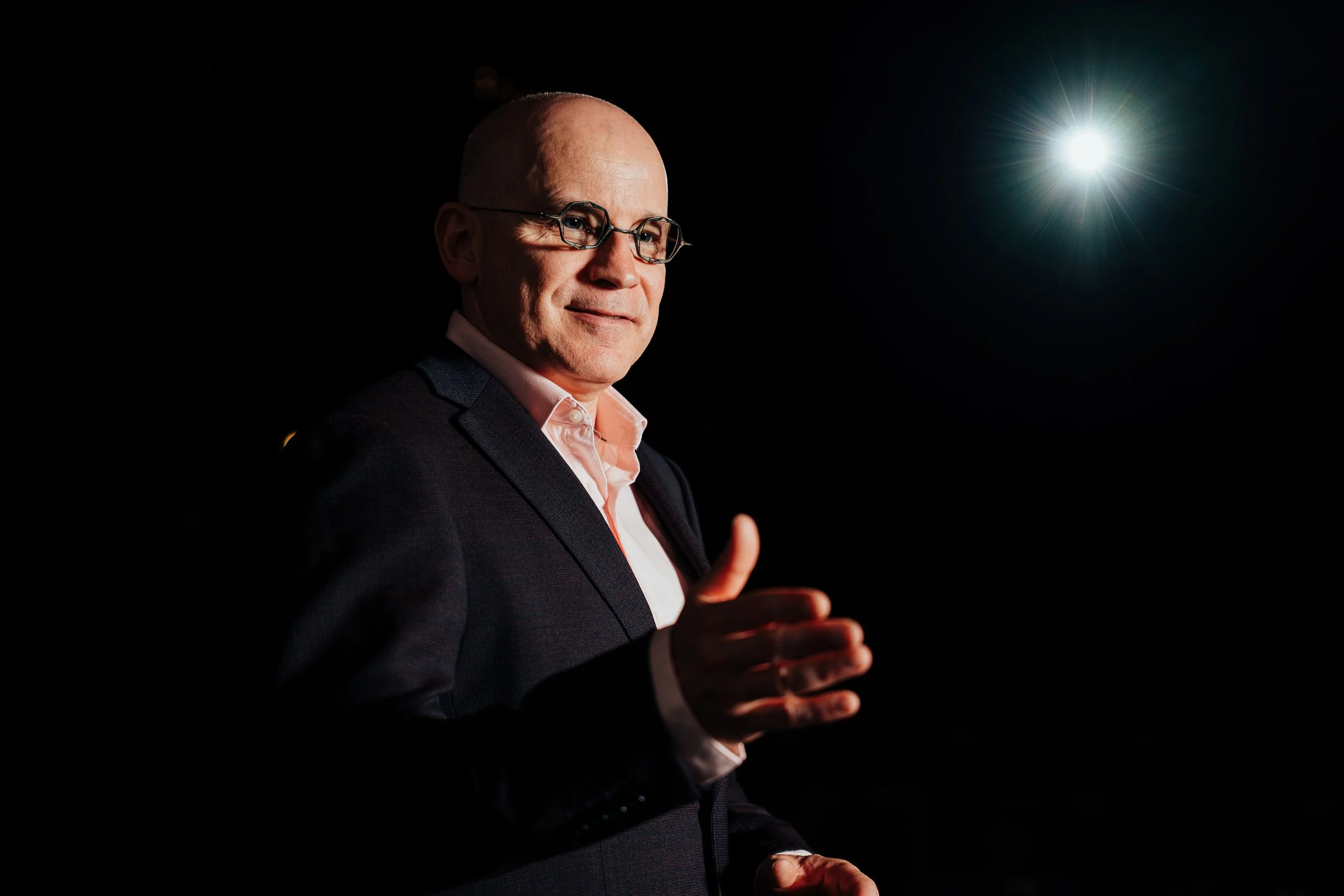 Homme portant des lunettes, souriant, en costume, avec un doigt levé, dans un environnement sombre avec une lumière brillante en arrière-plan.