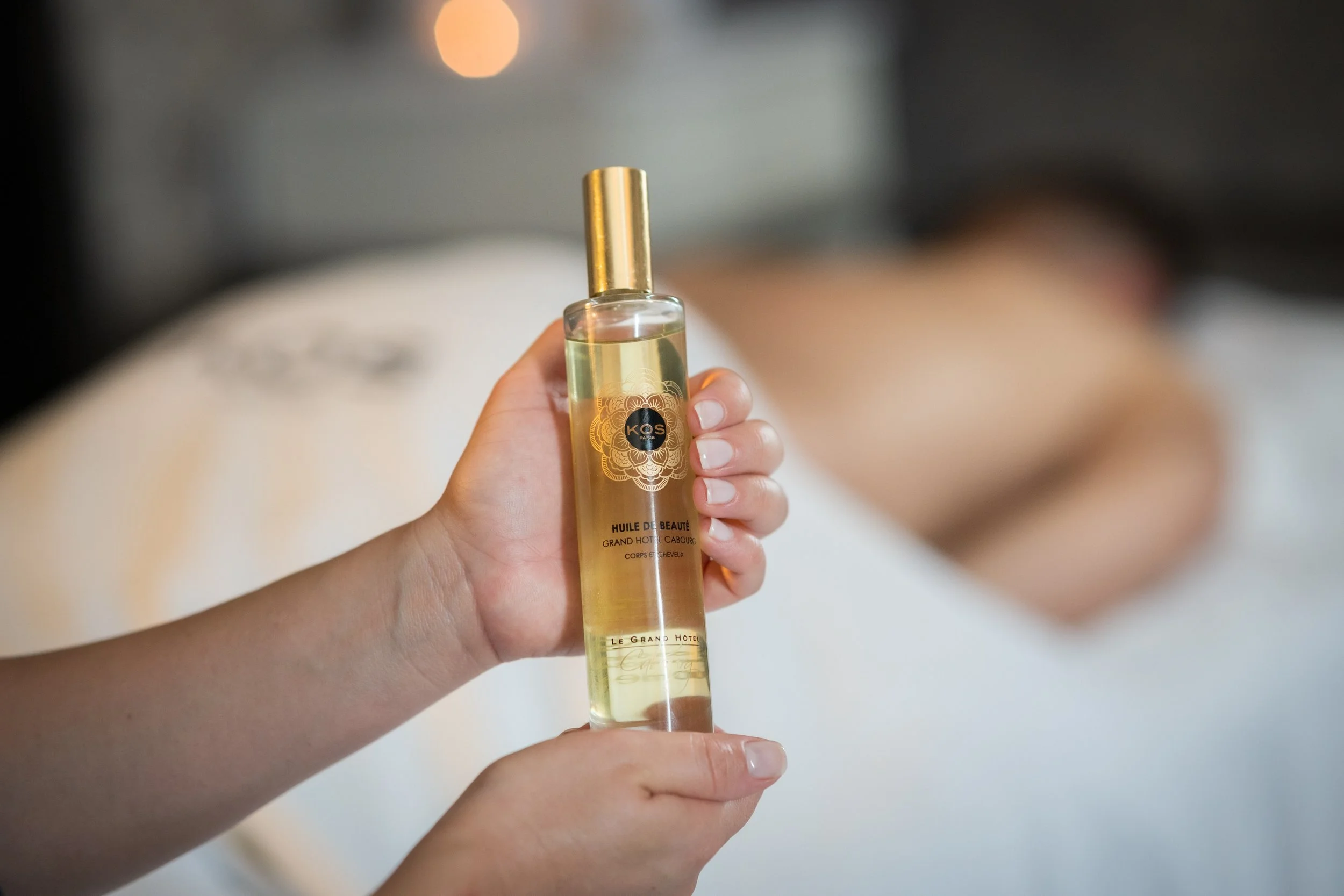 Une main tient un flacon en verre avec un liquide transparent, portant une étiquette dorée avec un motif floral et le texte "Huile de Beauté" et "Le Grand Hôtel Cabourg".
