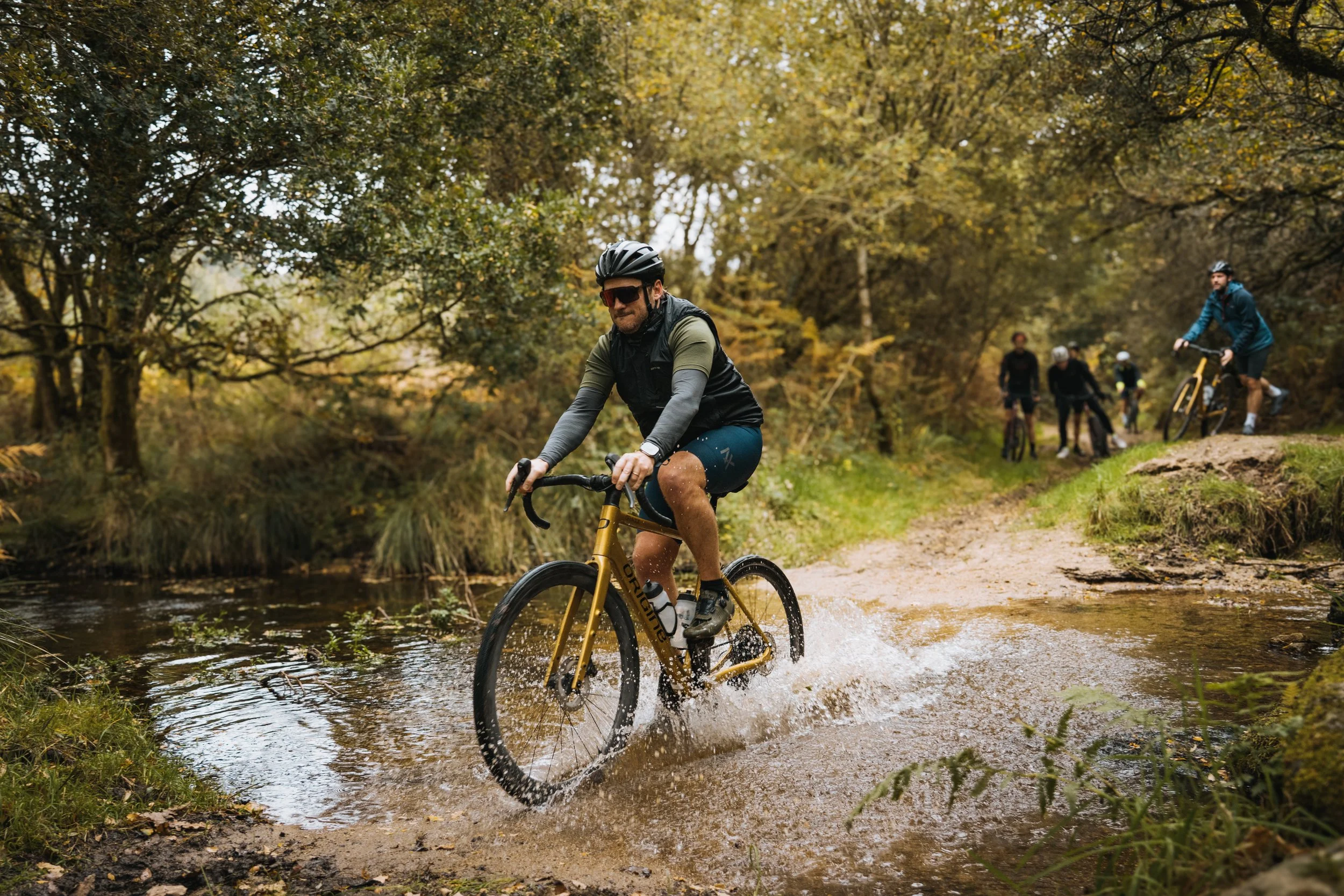 Groupe de personnes faisant du vélo en forêt lors d'une journée d'automne, certains traversant un ruisseau.