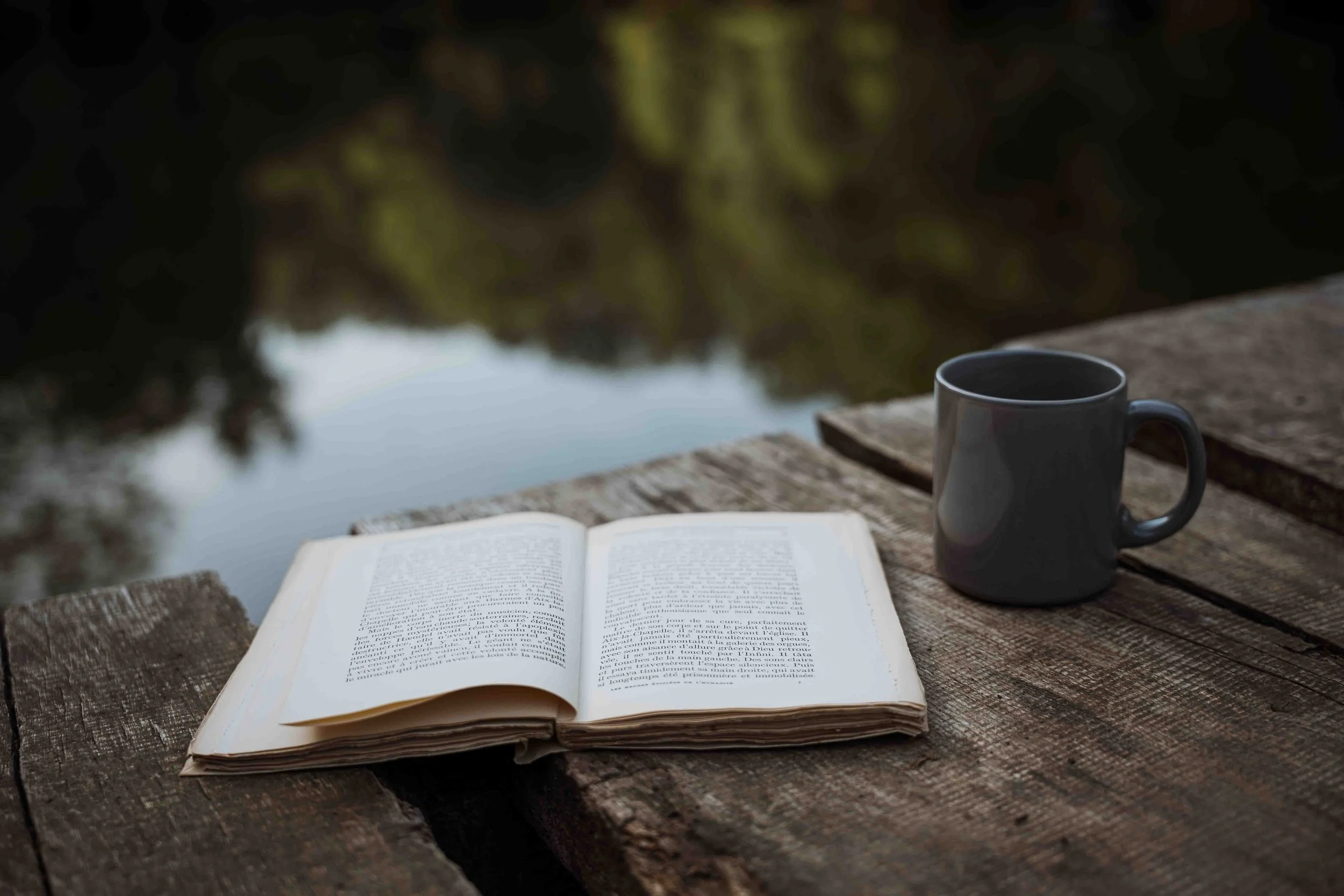 Un livre ouvert posé sur une table en bois à côté d'une tasse noire, avec un fond flou montrant un étang ou un lac