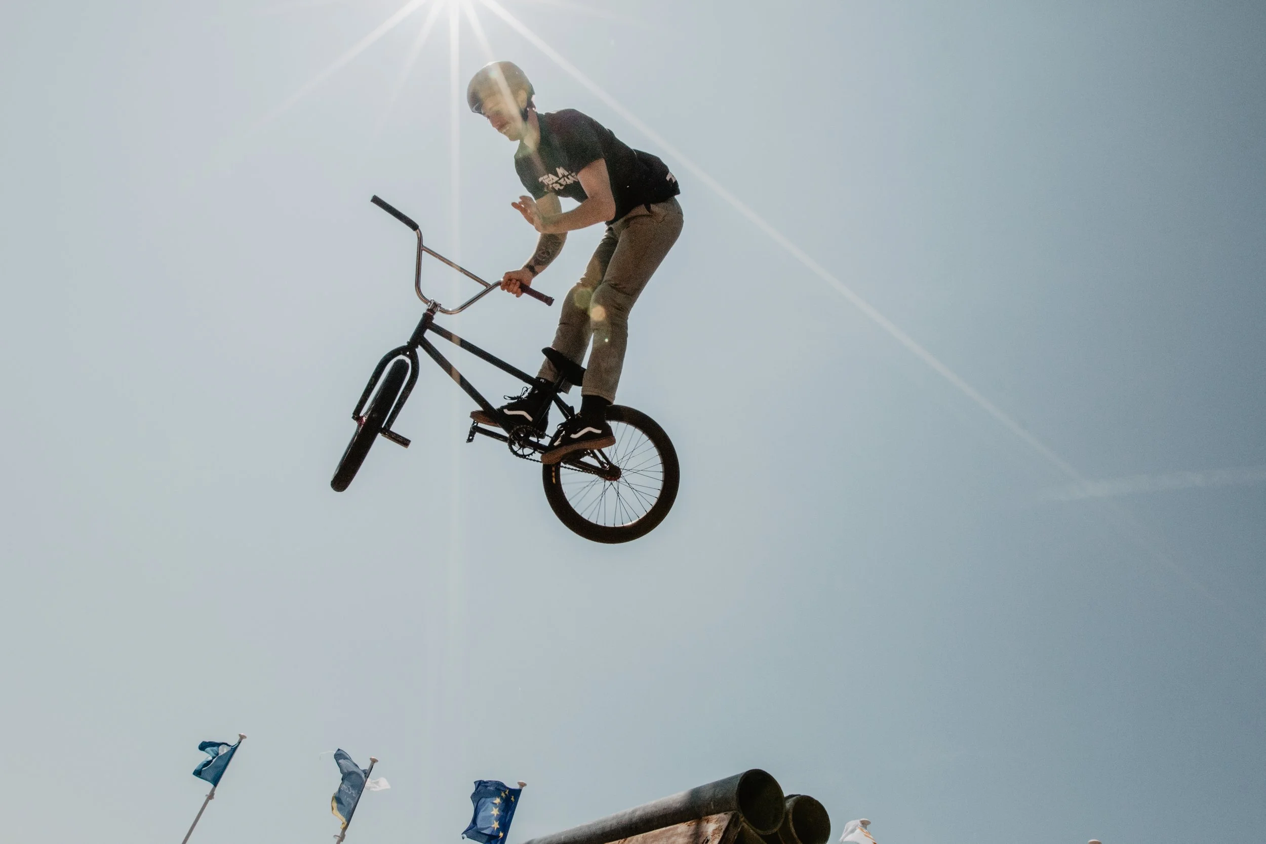 Une personne faisant une acrobatie aérienne sur un BMX en plein air sous un ciel dégagé ensoleillé, avec des drapeaux au premier plan.