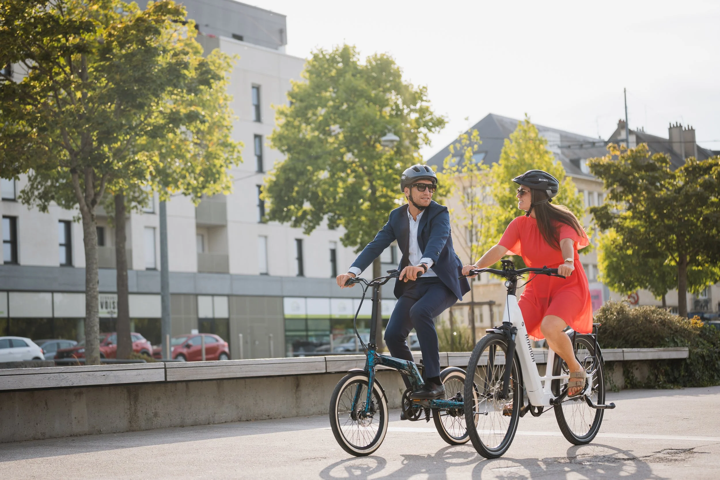 Un homme en costume et une femme en robe rouge, tous deux portant des casques de vélo, discutent en faisant du vélo dans un parc urbain ensoleillé avec des arbres et des bâtiments modernes en arrière-plan.