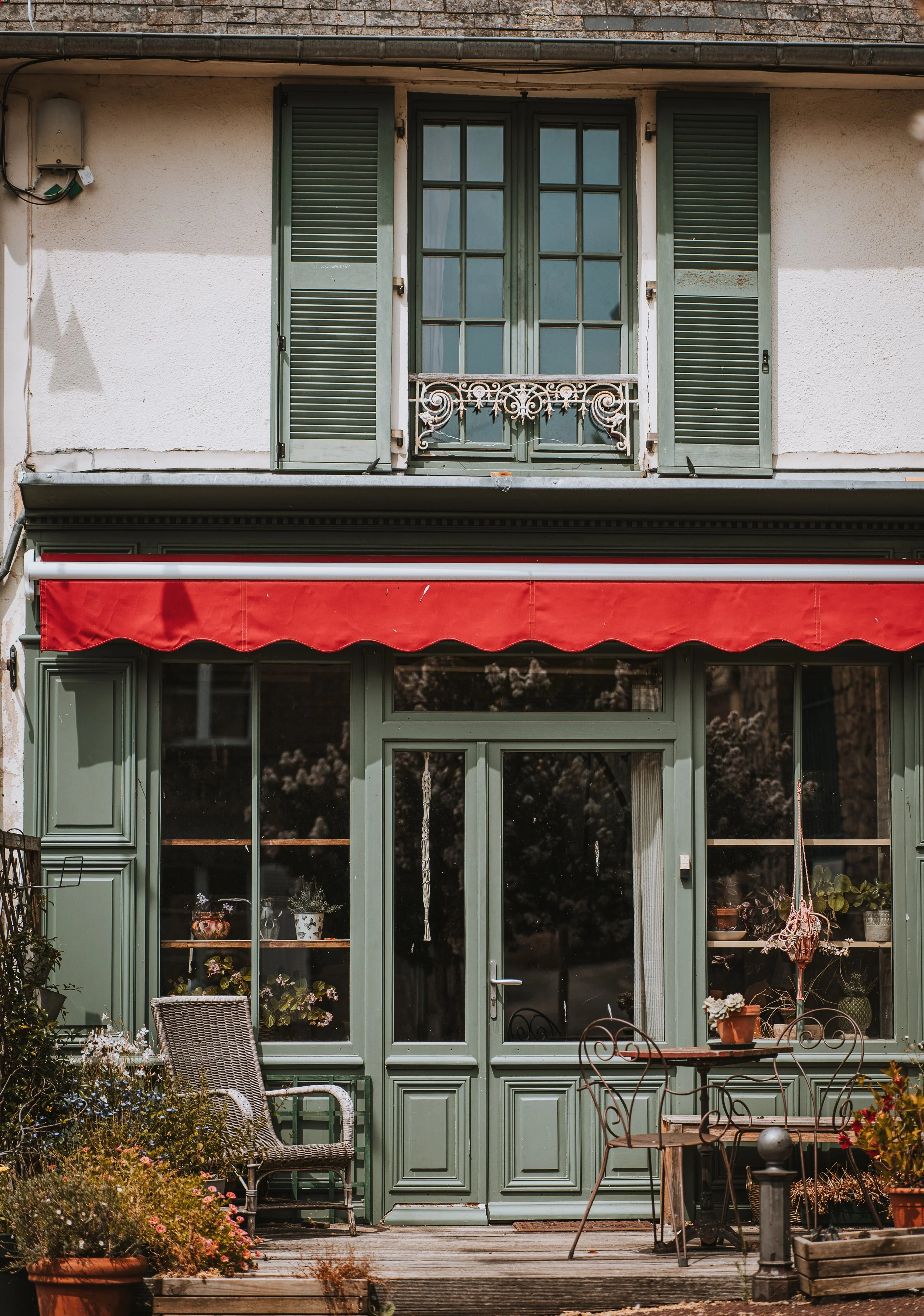 Une façade de maison avec fenêtres à volets verts, une porte en verre, et un balcon en fer forgé. Il y a des meubles d'extérieur, des plantes en pot, et une pergola avec un rideau rouge.