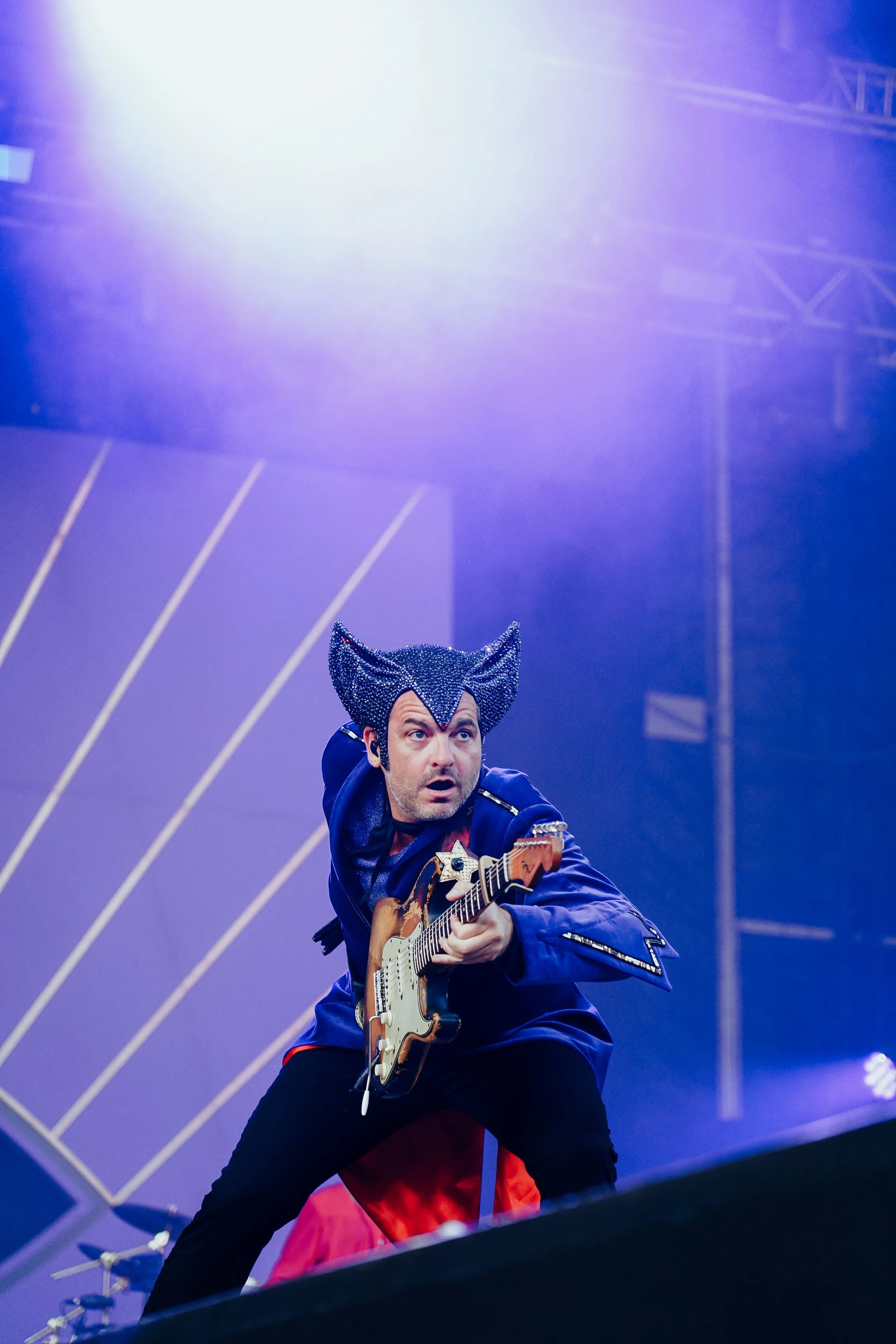 Un homme joue de la guitare sur scène, portant un chapeau en forme de chat noir pailleté, dans un éclairage violet et bleu.