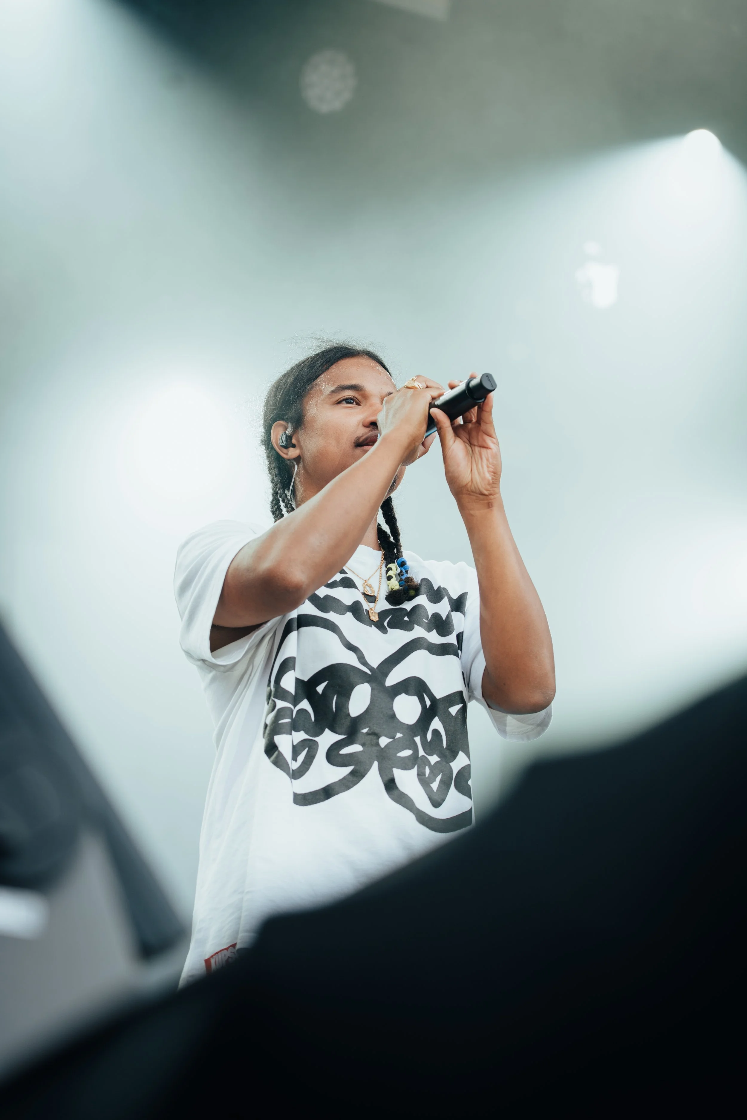 Une personne tenant un microphone lors d'une performance sur scène, portant un t-shirt avec un dessin de squelette et des boucles d'oreilles, éclairée par des lumières de scène.