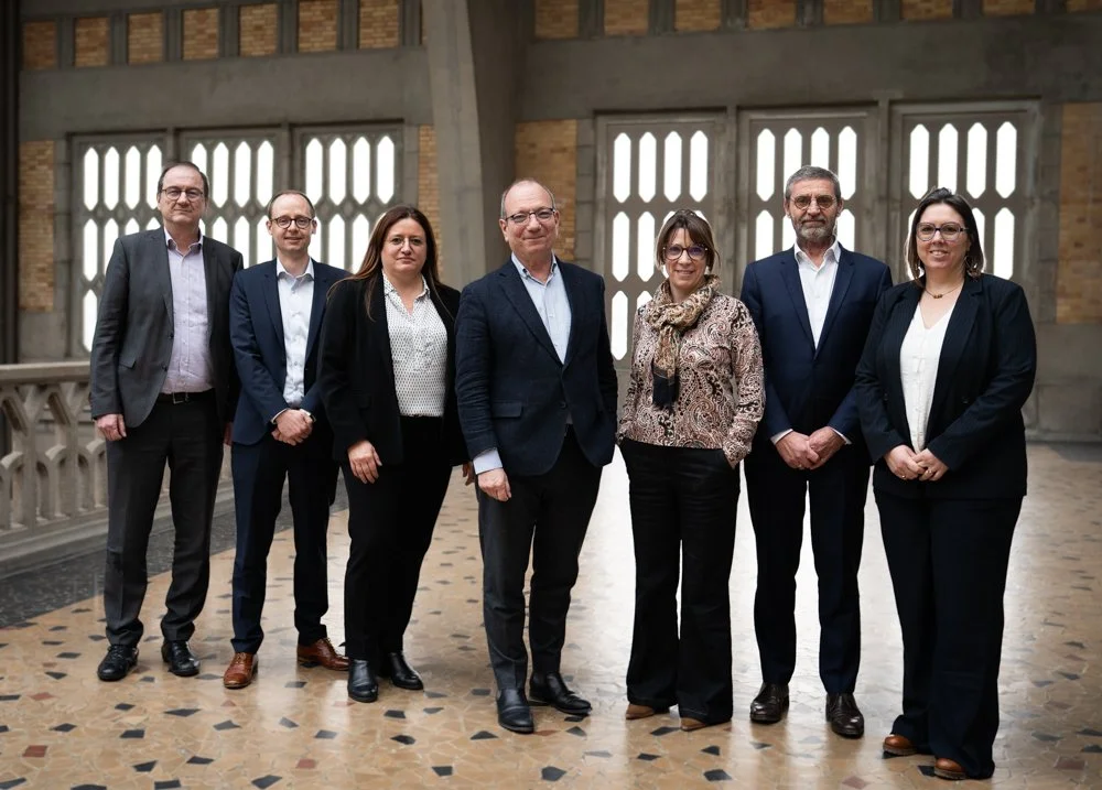 Un groupe de huit personnes en vêtements professionnels posant dans un bâtiment avec de grandes fenêtres en arrière-plan.