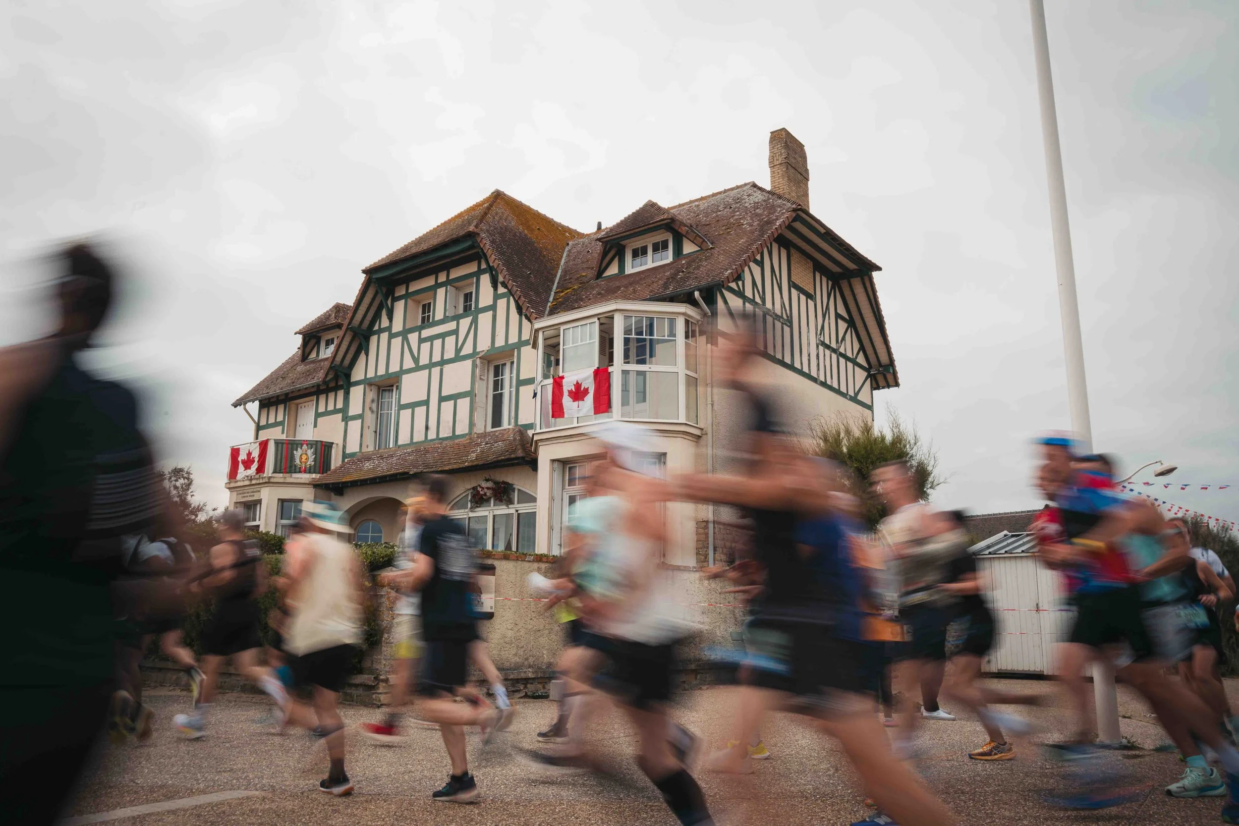 Une course ou un événement sportif avec de nombreux coureurs flous en mouvement devant une maison de style architectural traditionnel québécois, avec drapeaux canadiens et autres décorations.