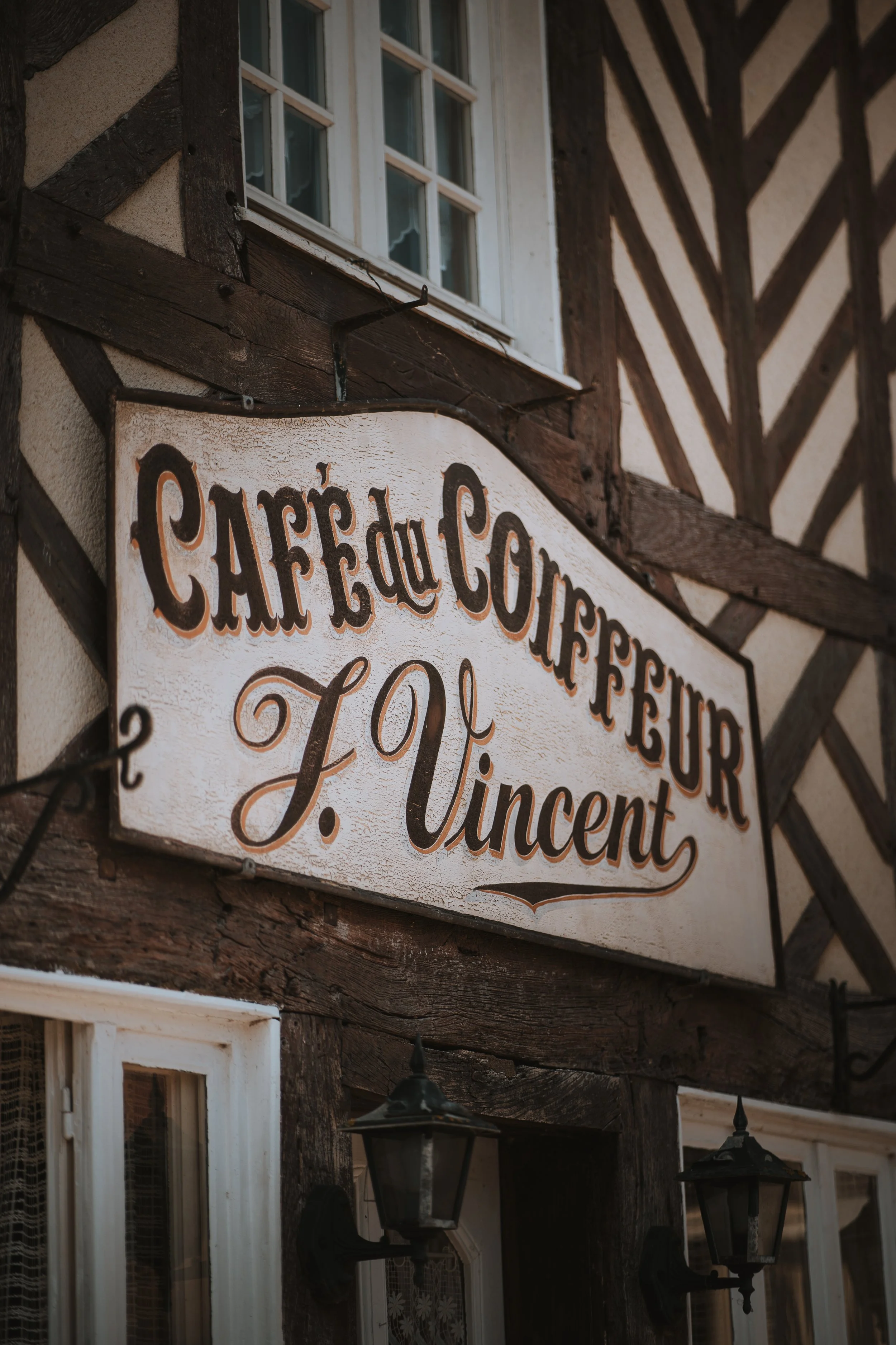 Une enseigne en bois fixée à une façade de bâtiment en style maison à colombages, indiquant un café, un café-concert, une salle de concert, avec une fenêtre à volets blancs en dessous.