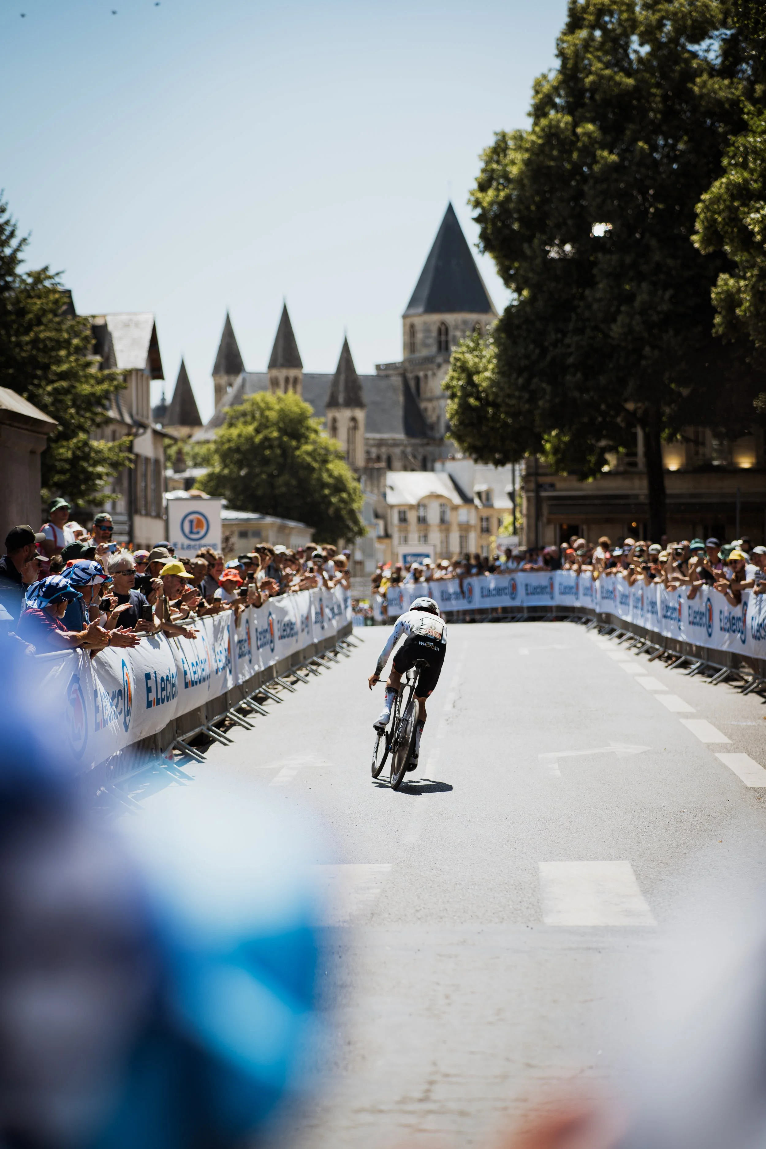 Un cycliste solo lors d'une course, vu à travers une ouverture ou un objet flou au premier plan. La foule et des bâtiments historiques sont visibles en arrière-plan.