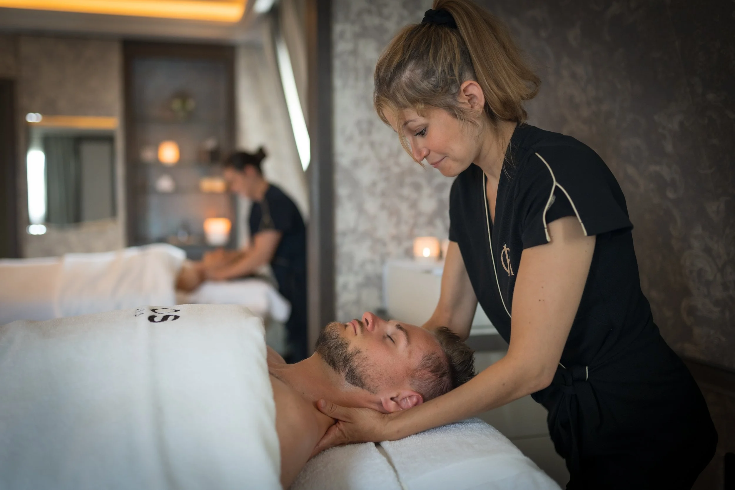 Une femme donne un massage à un homme allongé sur une table de massage dans une pièce relaxante, avec une autre femme en arrière-plan.