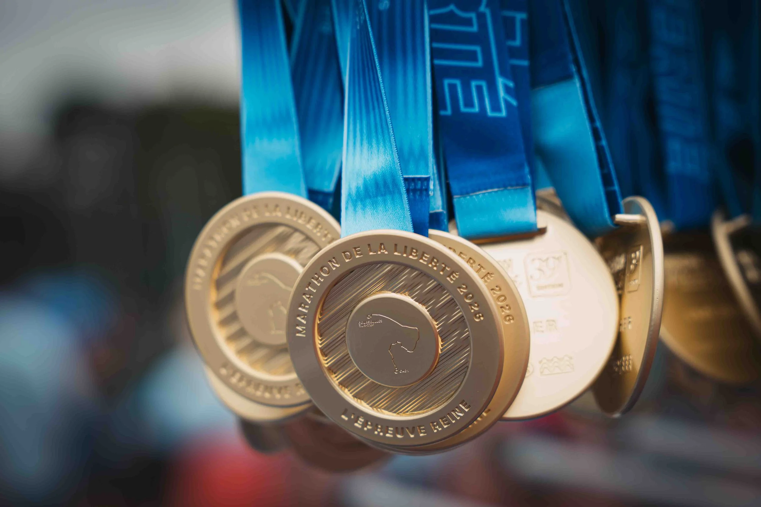 Des médailles d'or avec un ruban bleu suspendues, portant des inscriptions en français comme 'LAUREAT DE LA LIBERTÉ' et '2026'.
