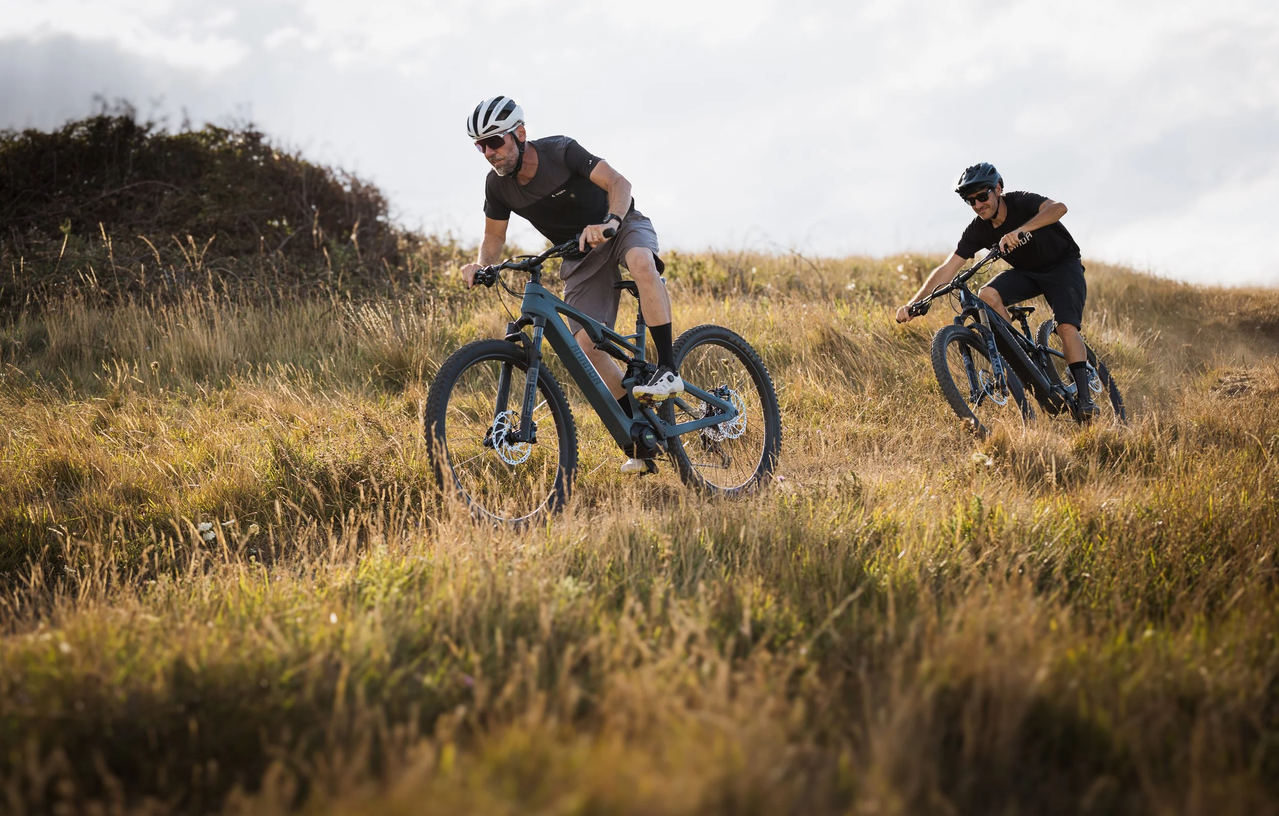 Deux hommes faisant du vélo sur un terrain herbeux en plein air, portant des casques de protection et des vêtements décontractés, avec un ciel partiellement nuageux en arrière-plan.