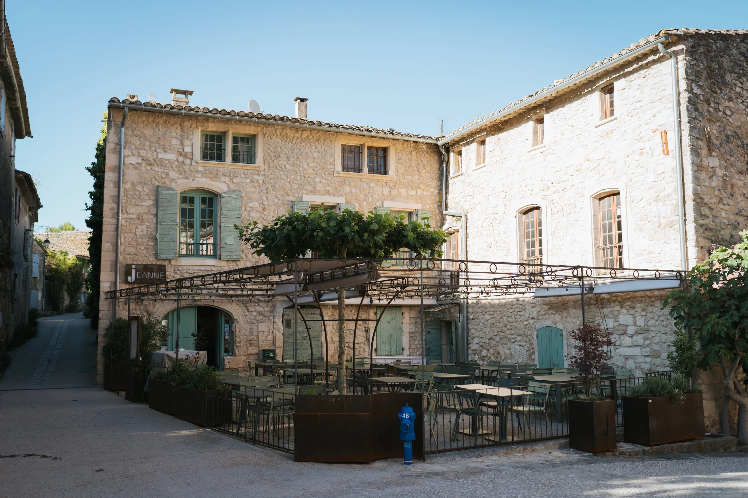 Un bâtiment en pierre avec des fenêtres à volets verts et une terrasse extérieure avec des chaises et des tables, entouré d'un mur en fer et de plantes, sous un ciel bleu clair.