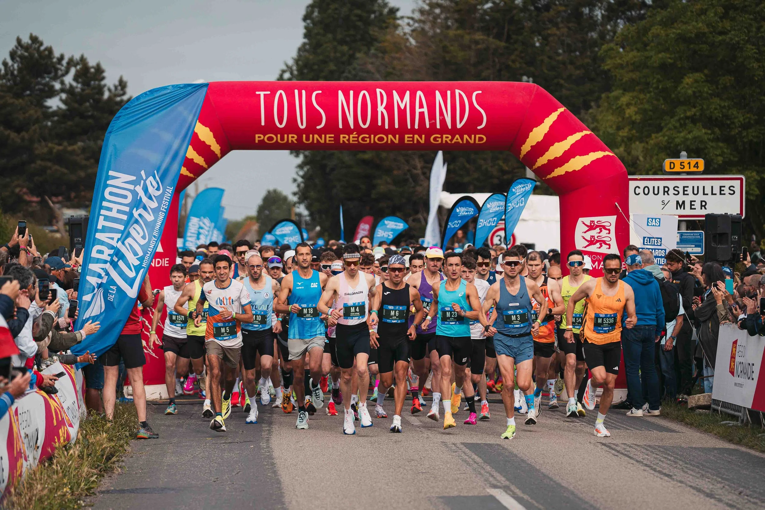 Départ d'une course de marathon avec de nombreux coureurs, un arch en forme de tunnel rouge avec écrit 'TOUS NORMANDS POUR UNE RÉGION EN GRAND' en haut, et des drapeaux bleus autour.