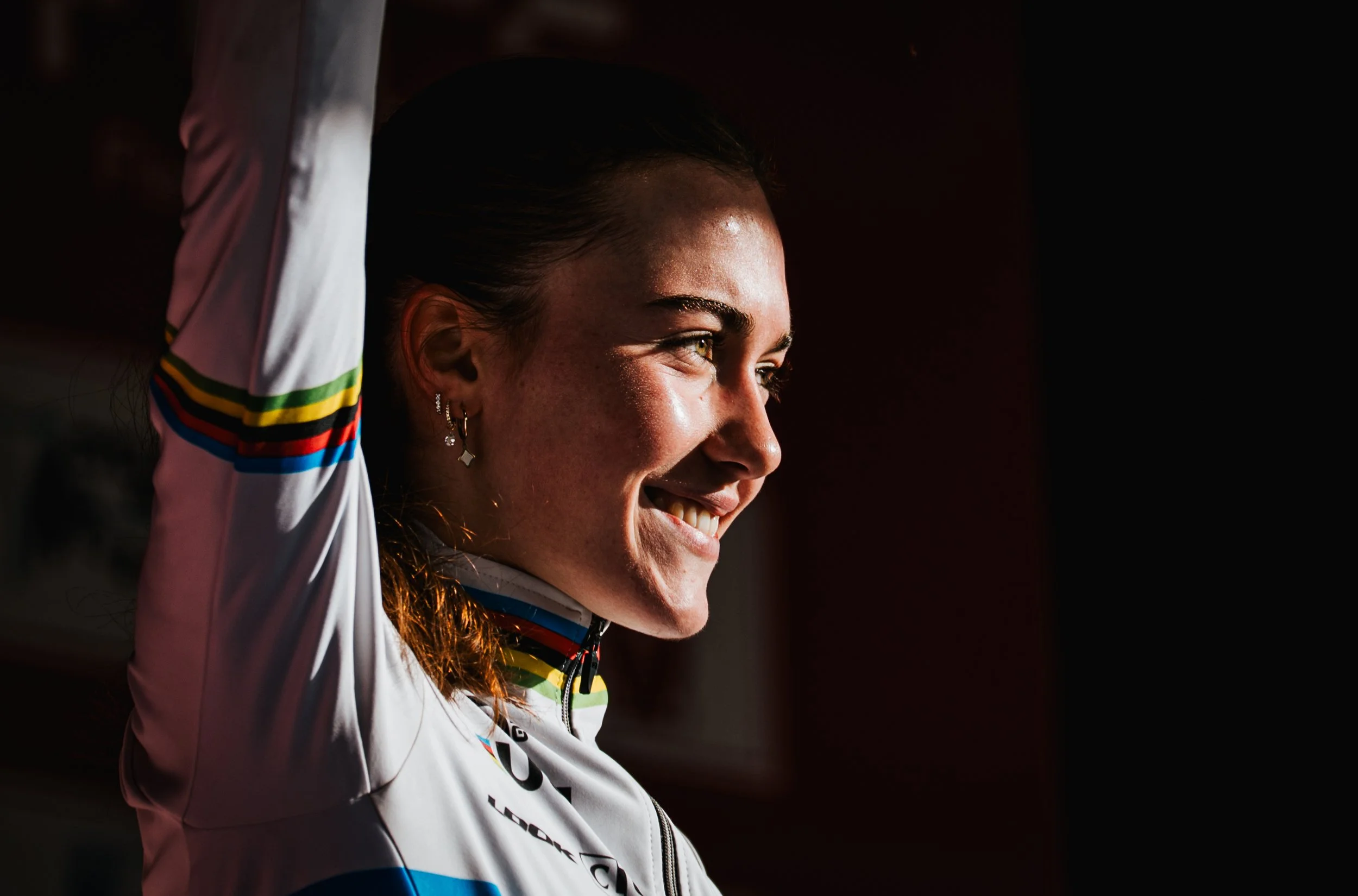 Une femme souriante avec des earrings, portant une veste de sport blanche avec des bandes colorées, observant vers la droite dans un éclairage chaud.