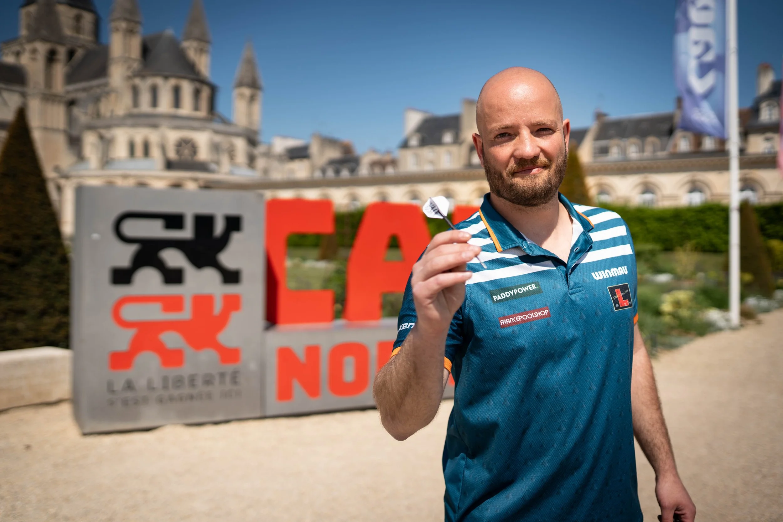 Un homme avec une barbe portant un maillot de sport bleu et blanc tient une fléchette devant une grande affiche avec le mot 'STOP' en rouge. En arrière-plan, un château et un ciel bleu clair.