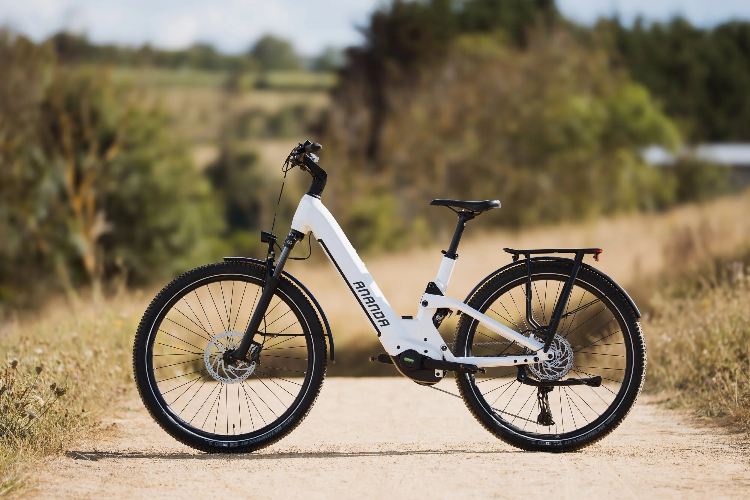 Vélo électrique blanc avec cadre épuré, pneus épais et sac à l'arrière, stationné sur un sentier naturel entouré d'herbes et d'arbres, sous un ciel clair.