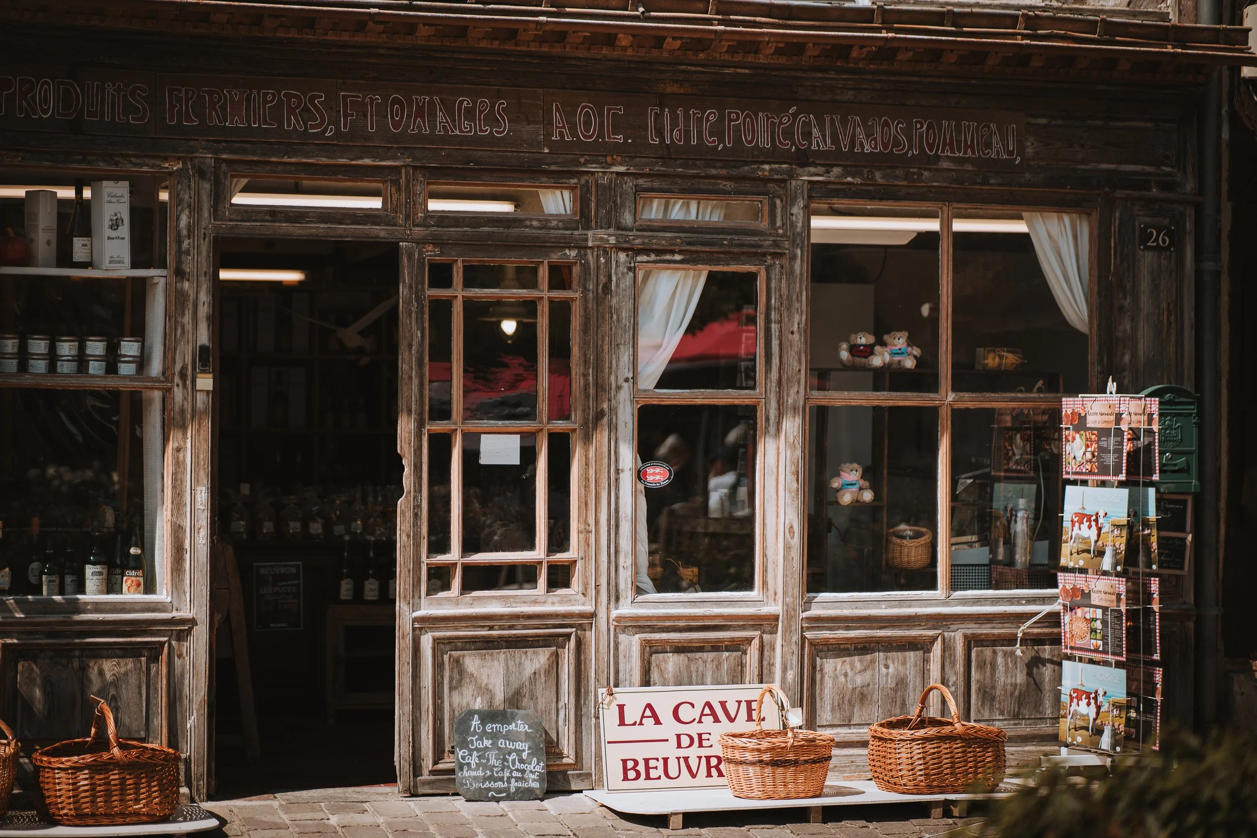 Une façade de boutique en bois avec fenêtres en verre et panneau en bois écrit en français, vendant des produits locaux, fromage, et produits du terroir, décorée avec des paniers en osier à l'extérieur.