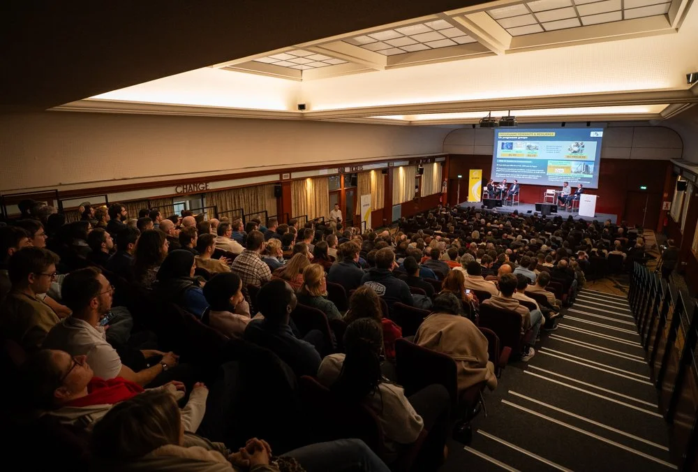 Une grande salle de conférence remplie de personnes assises, regardant un panneau d'affichage numérique où une présentation est en cours.
