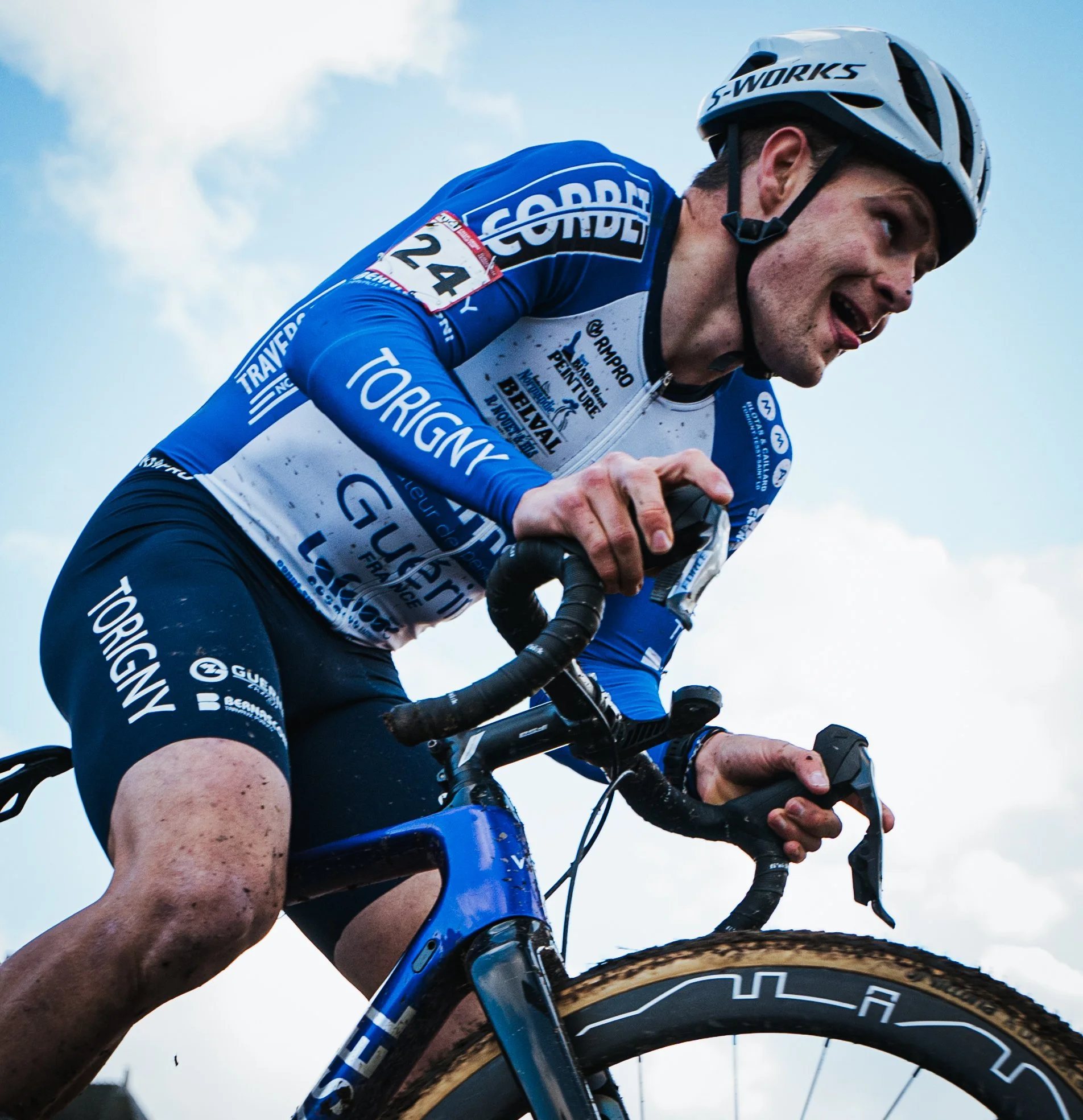 Cycliste en pleine course, portant un casque blanc et un maillot bleu avec divers logos, chevauche un vélo noir avec des roues en bois, sous un ciel partiellement nuageux.