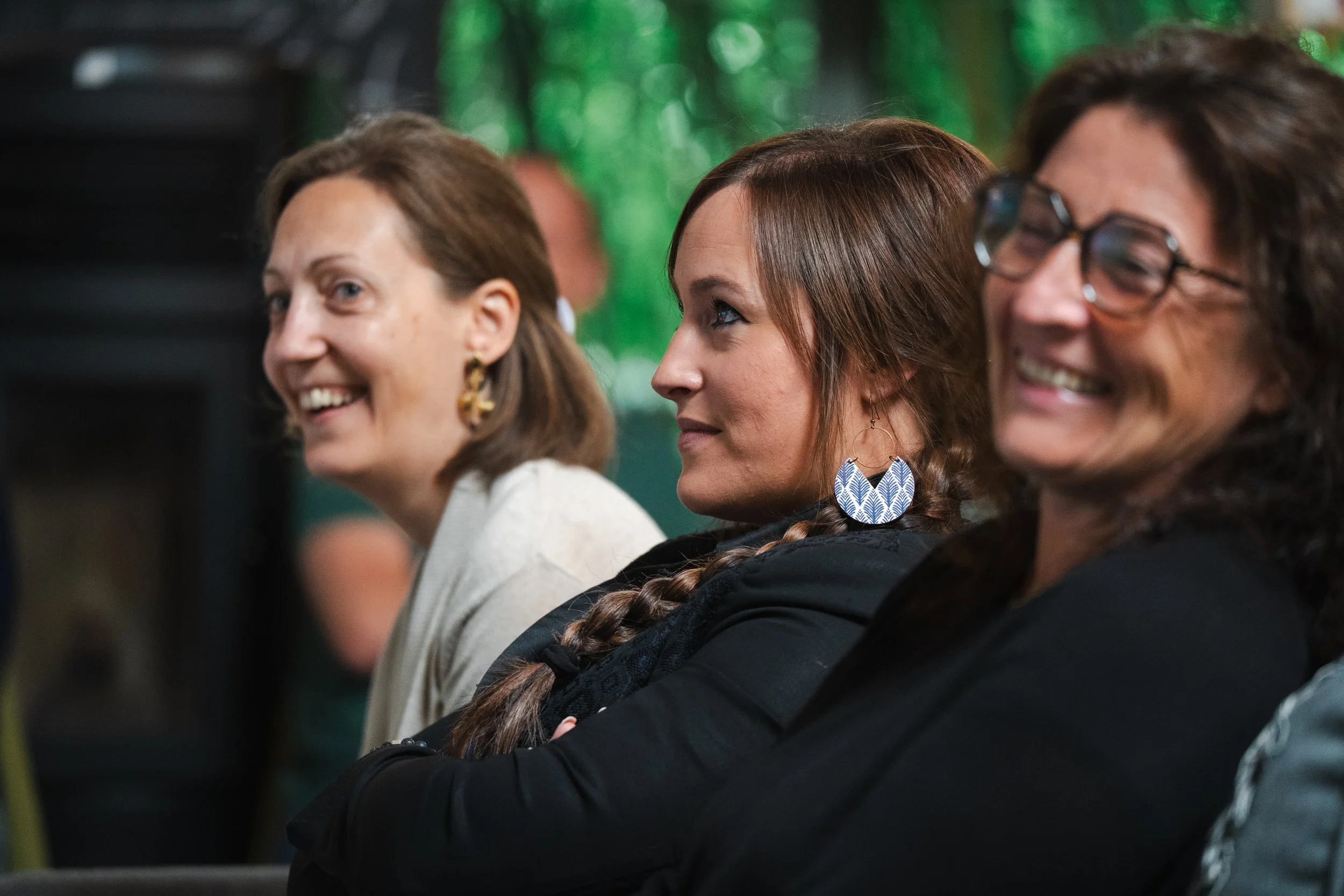 Trois femmes souriantes assises de profil lors d'une réunion ou d'événement, avec un fond flou de verdure ou d'éclairage naturel.