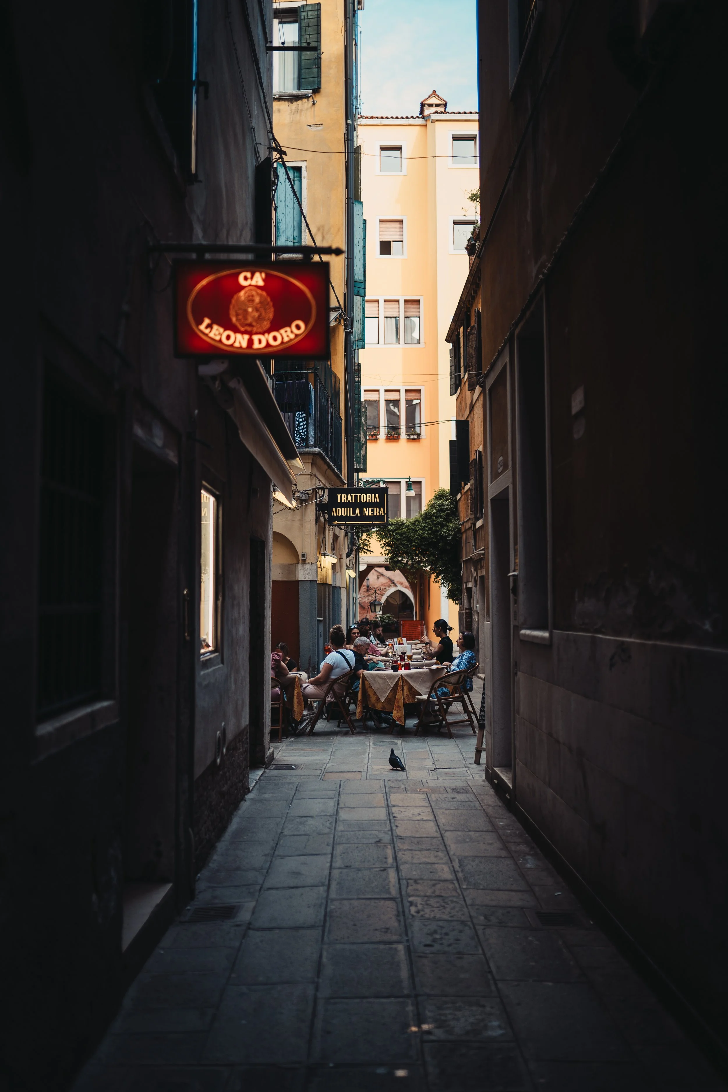 Une ruelle étroite dans une ville européenne avec des terrasses de cafés en plein air où des gens dégustent des repas. Au fond, des bâtiments aux fenêtres avec volets en bois. Il y a une enseigne lumineuse rouge avec le texte 'Cà Leone D'Oro' et une autre plaque noire indiquant 'Trattoria Aquila Nera'.