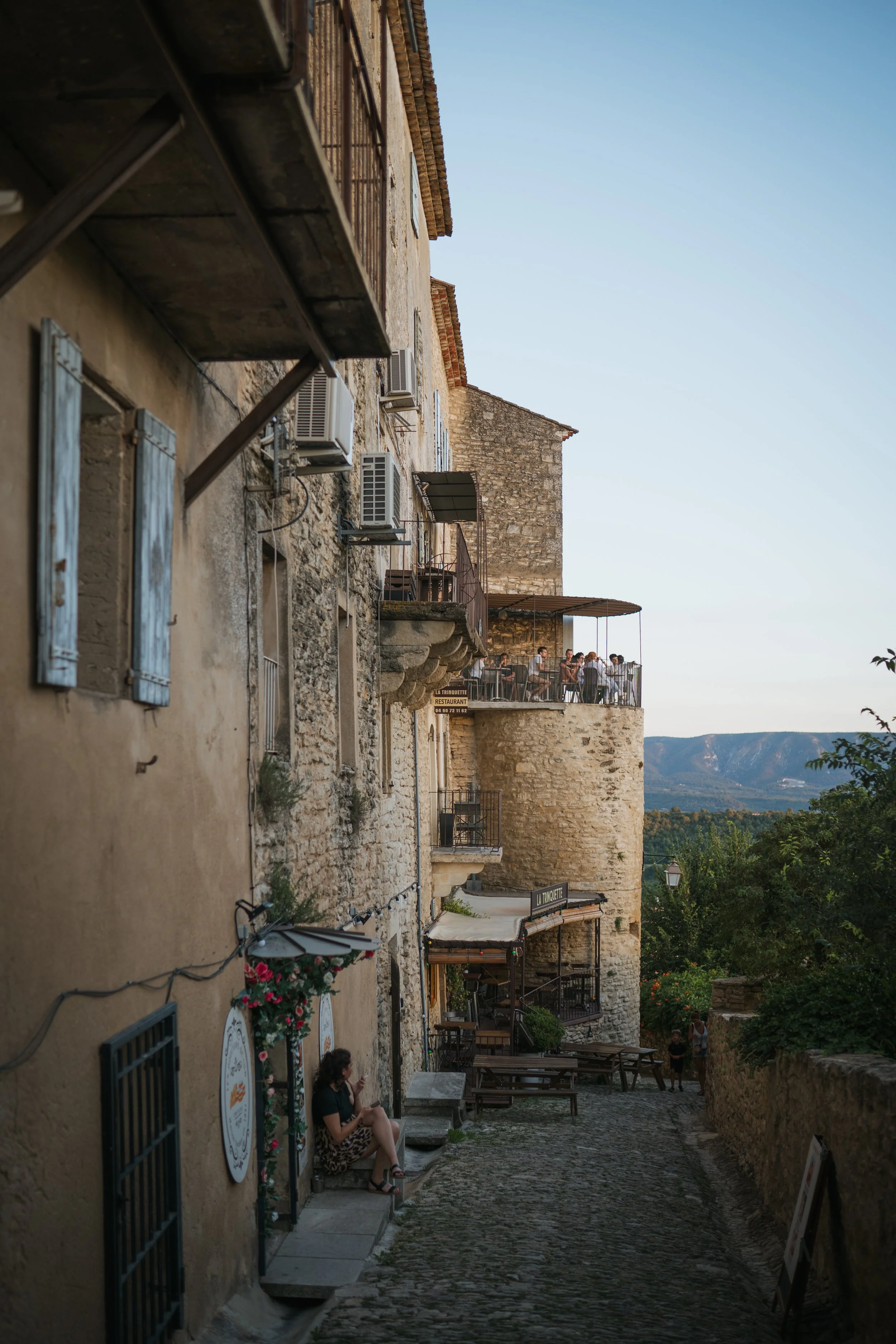 Une rue pavée dans un village avec des bâtiments en pierre, des volets bleus, et des terrasses avec des personnes assises, un paysage de montagnes au loin, sous un ciel clair.