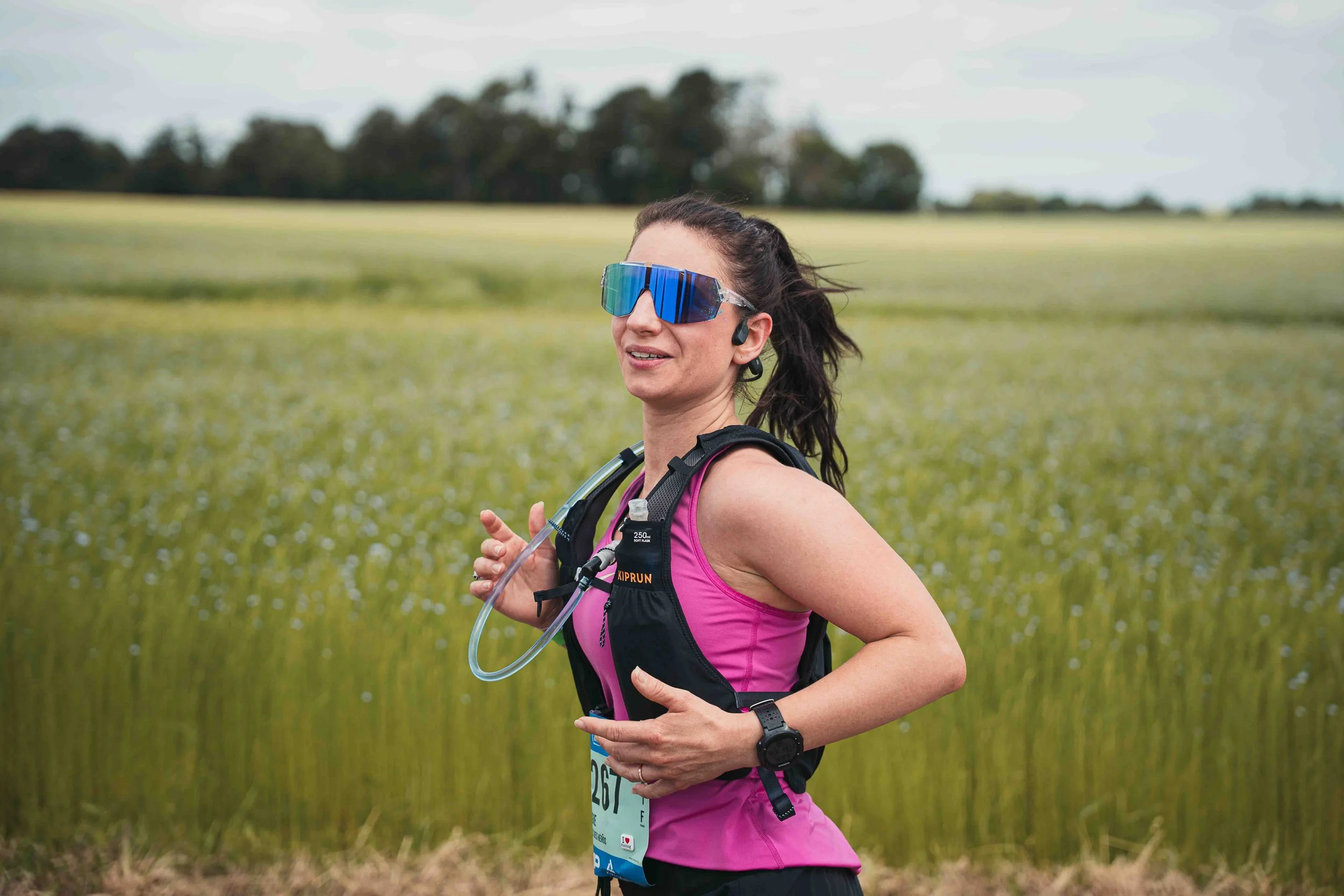 Une femme court dans un champ herbeux, portant des lunettes de soleil sportives, un vêtement de course rose et un harnais avec une gourde, en pleine nature.