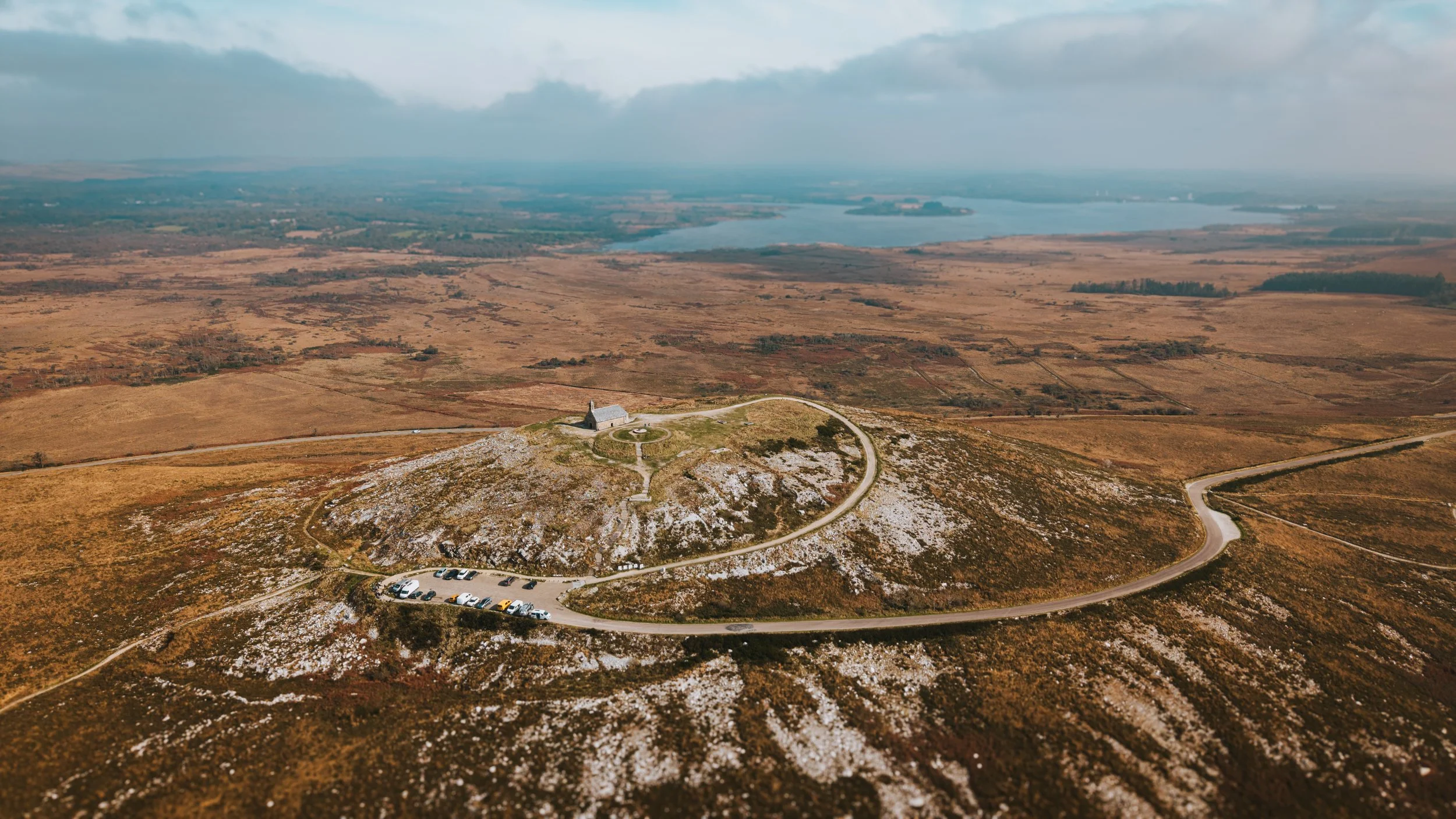 Une vue aérienne d'une colline avec une petite chapelle en pierre et un chemin en boucle, entourée d'une zone de terrain dégagé avec quelques voitures garées, au-dessus d'une plaine avec un lac et un ciel nuageux.
