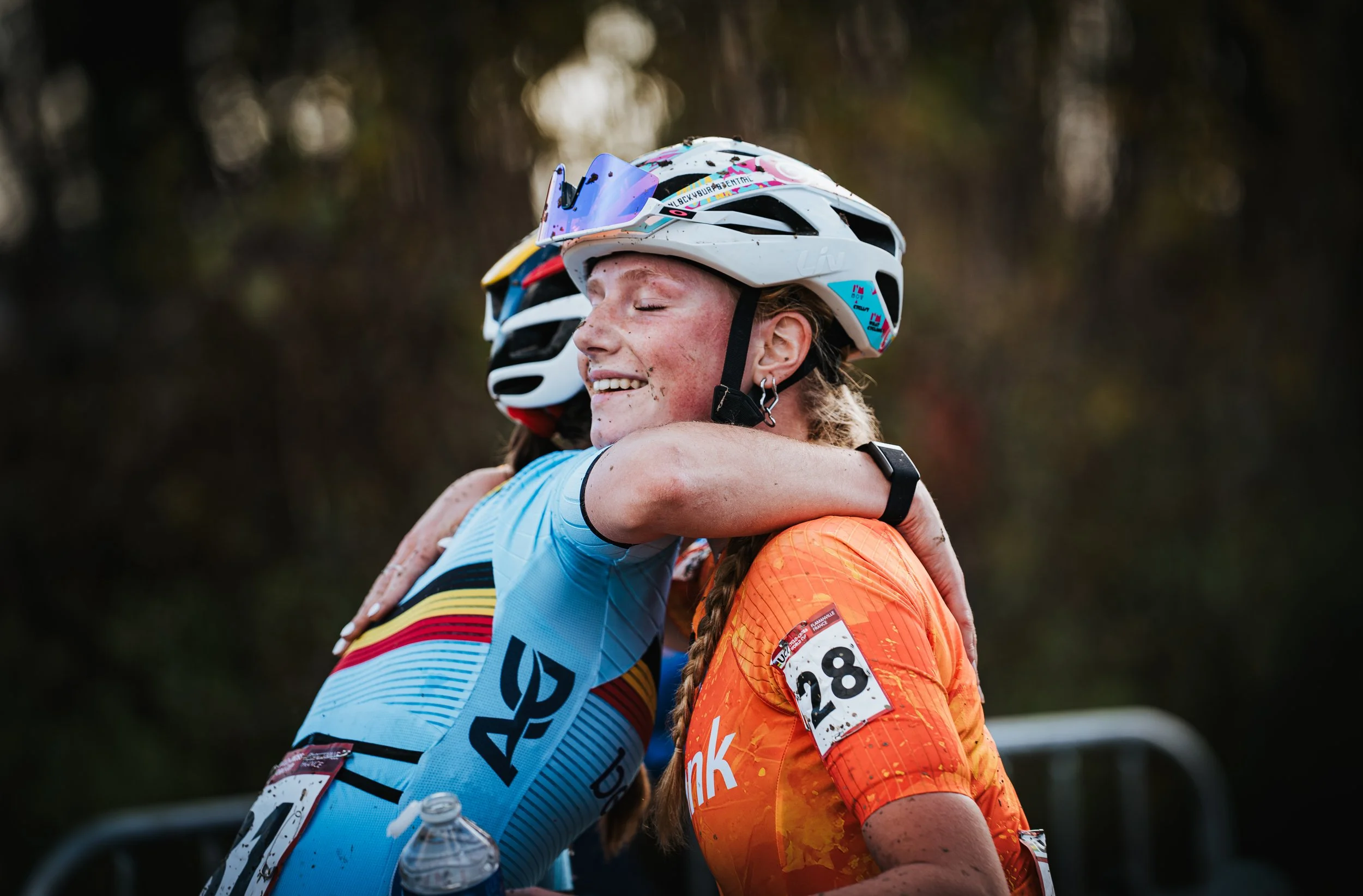 Deux femmes cyclistes en tenue de sport s'embrassent, portant des casques de vélo, dans une nature forestière.