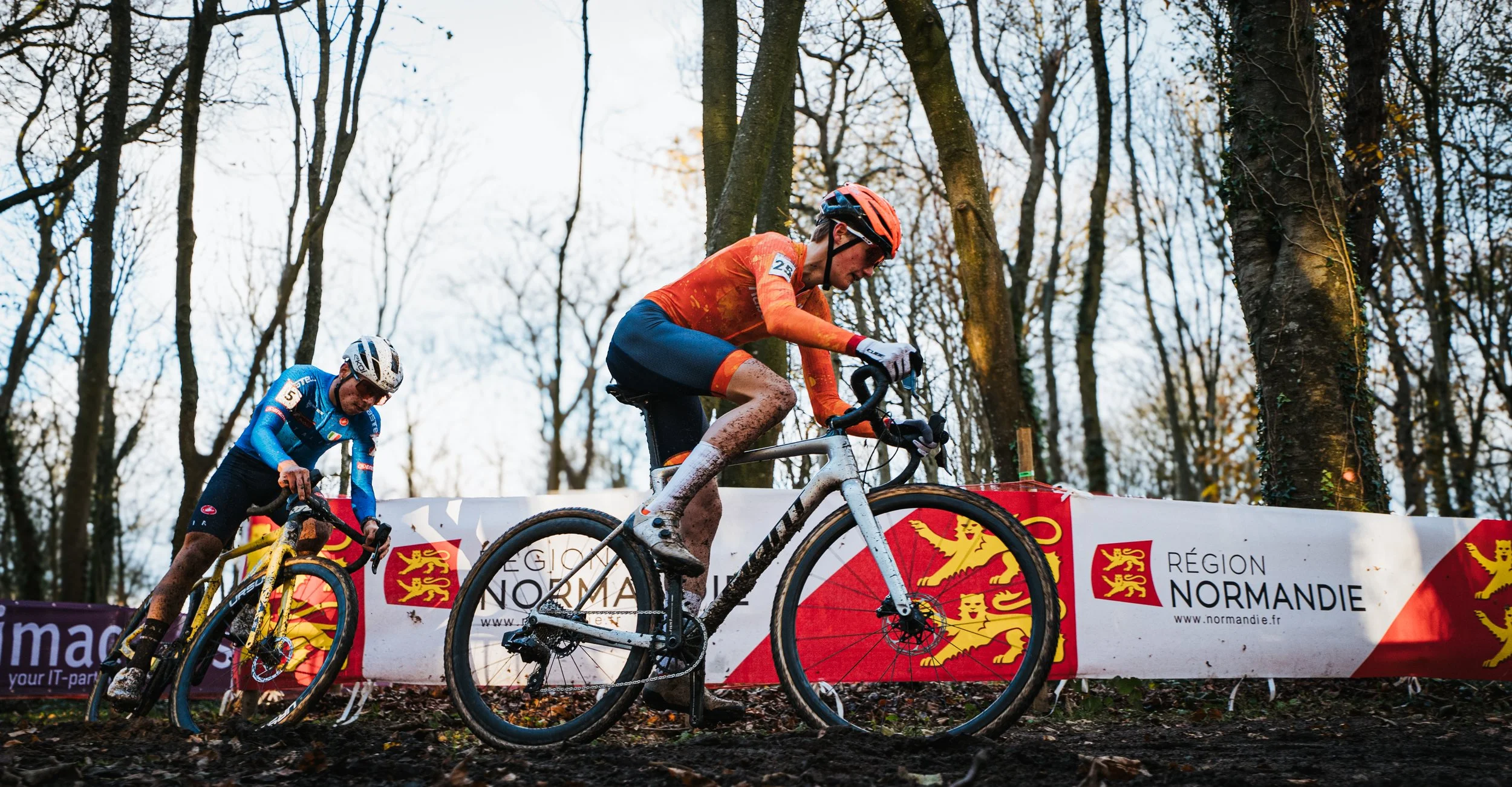 Deux cyclistes participant à une course en forêt, portant des tenues de sport colorées, dont un en orange et noir, avec un panneau publicitaire en arrière-plan indiquant la région Normandie.
