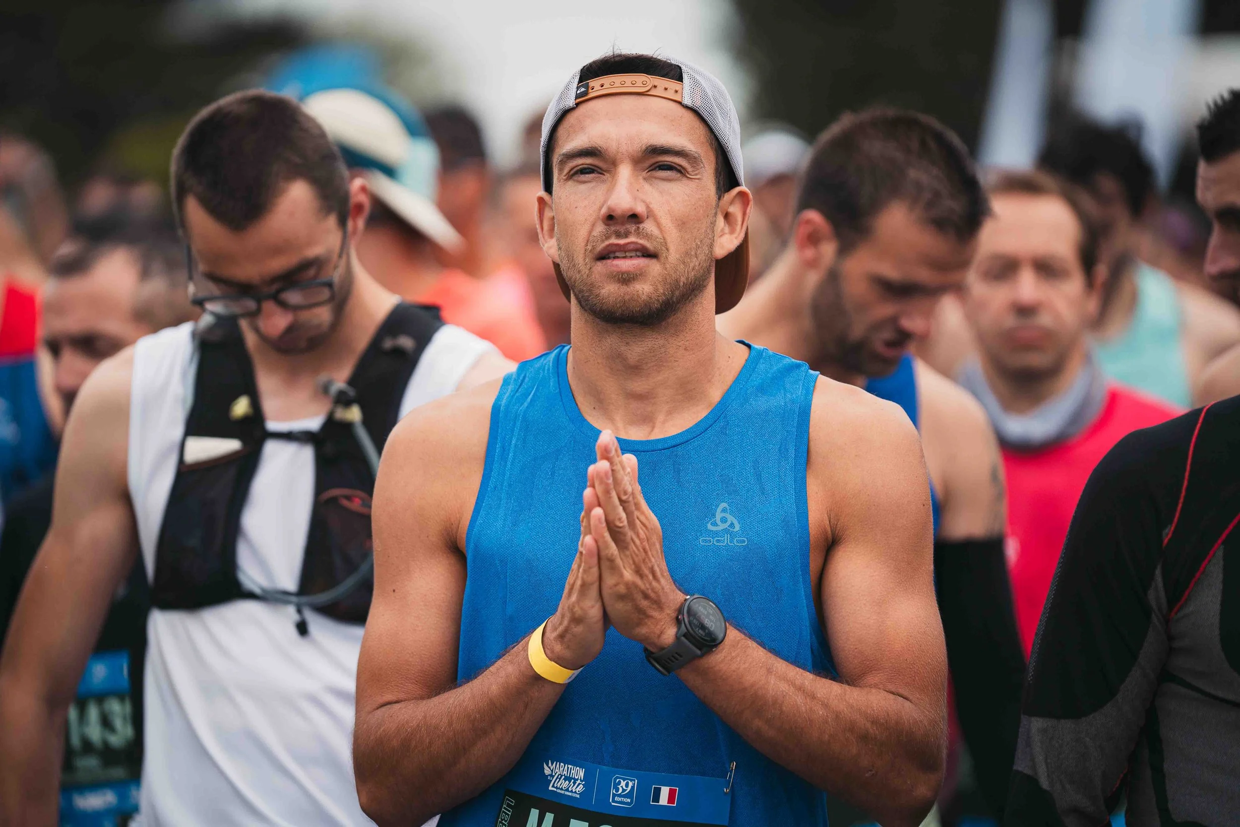 Un homme vêtu d'une chemise en nylon bleu, portant une montre et une ceinture, pratique la méditation lors d'une course ou d'un événement sportif, entouré d'autres coureurs.