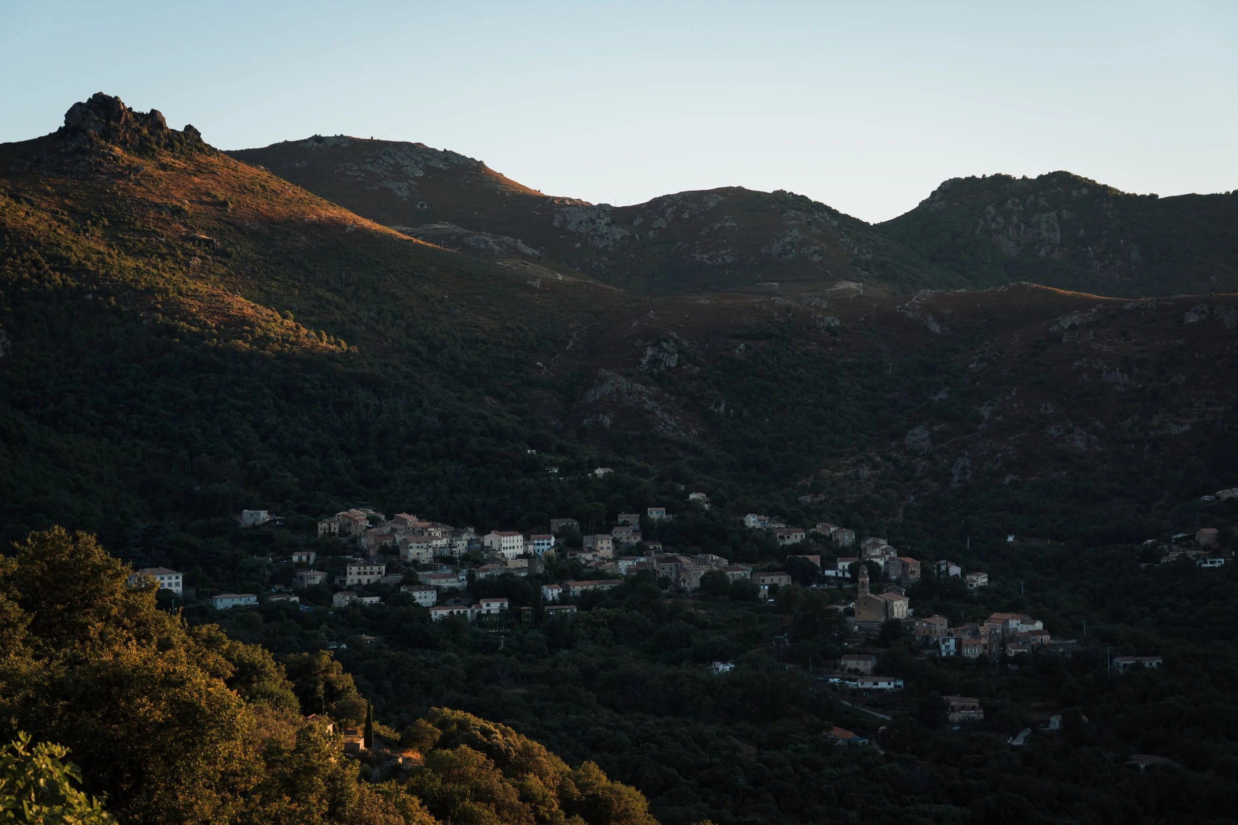 Village de petites maisons dans une vallée entourée de montagnes avec peu de végétation, au coucher du soleil.