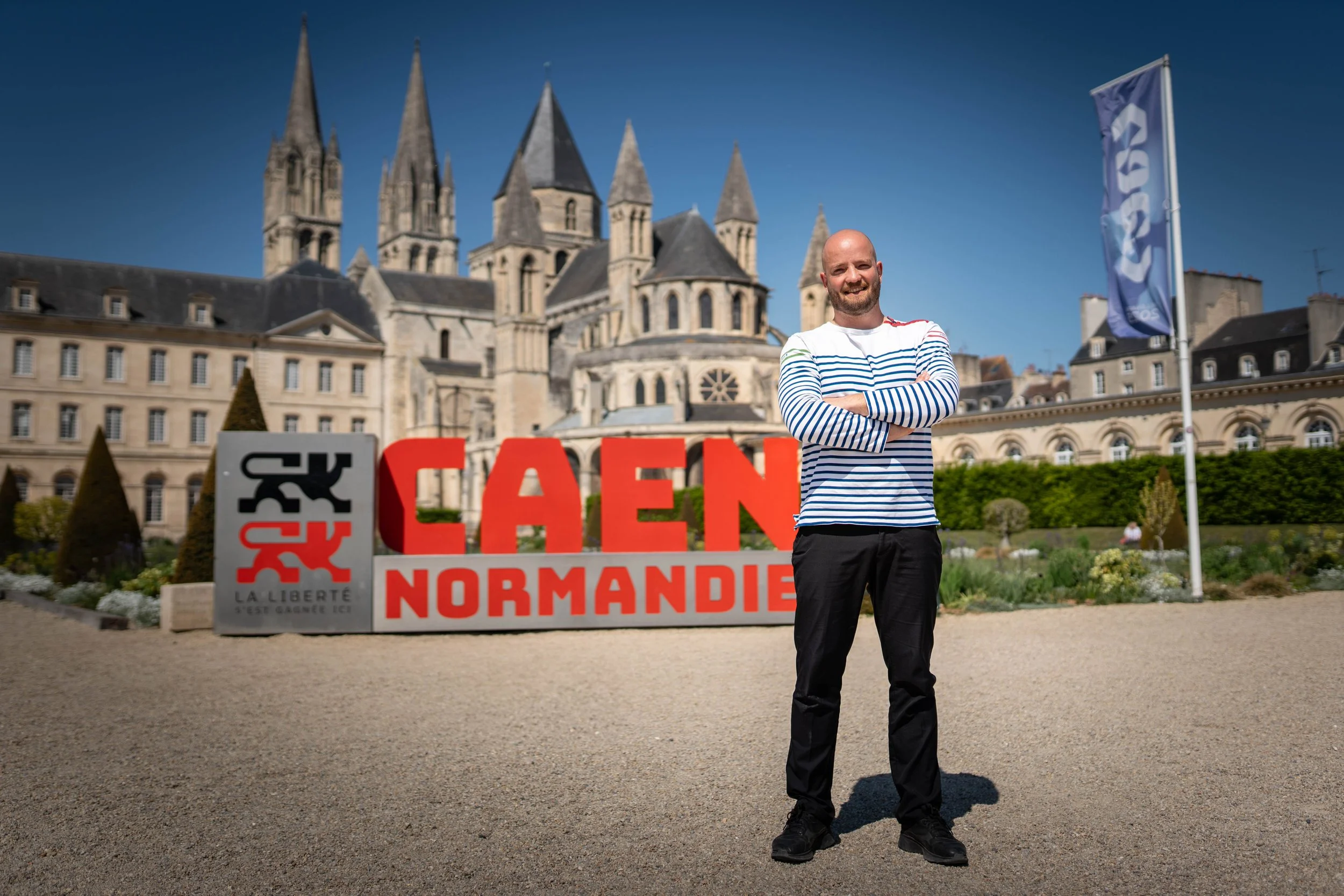 Un homme souriant en pull à rayures bleues et blanches et pantalons noirs, debout devant un panneau indiquant 'Normandie' avec un symbole de liberté, en arrière-plan une grande cathédrale en pierre et un drapeau bleu, dans un parc ou une place publique en Normandie.