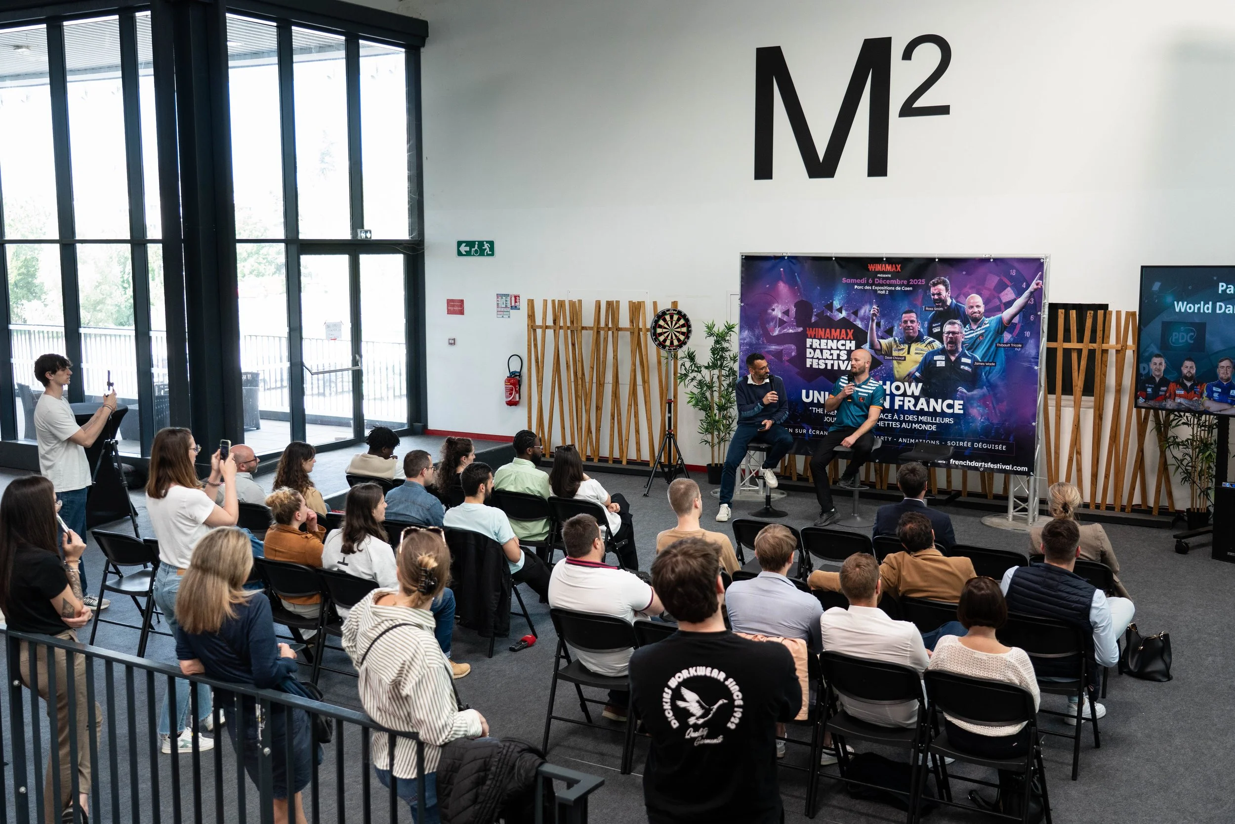 Audience assistant à une conférence ou un événement esport dans une grande salle intérieure avec un panneau d'affichage promotionnel pour le festival de fléchettes et un écran montrant des joueurs de sport électroniques.