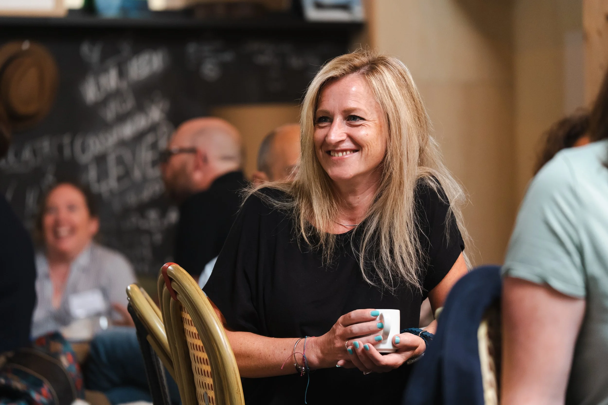 Une femme aux cheveux blonds et longs, souriante, tenant une tasse dans un environnement de réunion ou de café, entourée de plusieurs personnes.