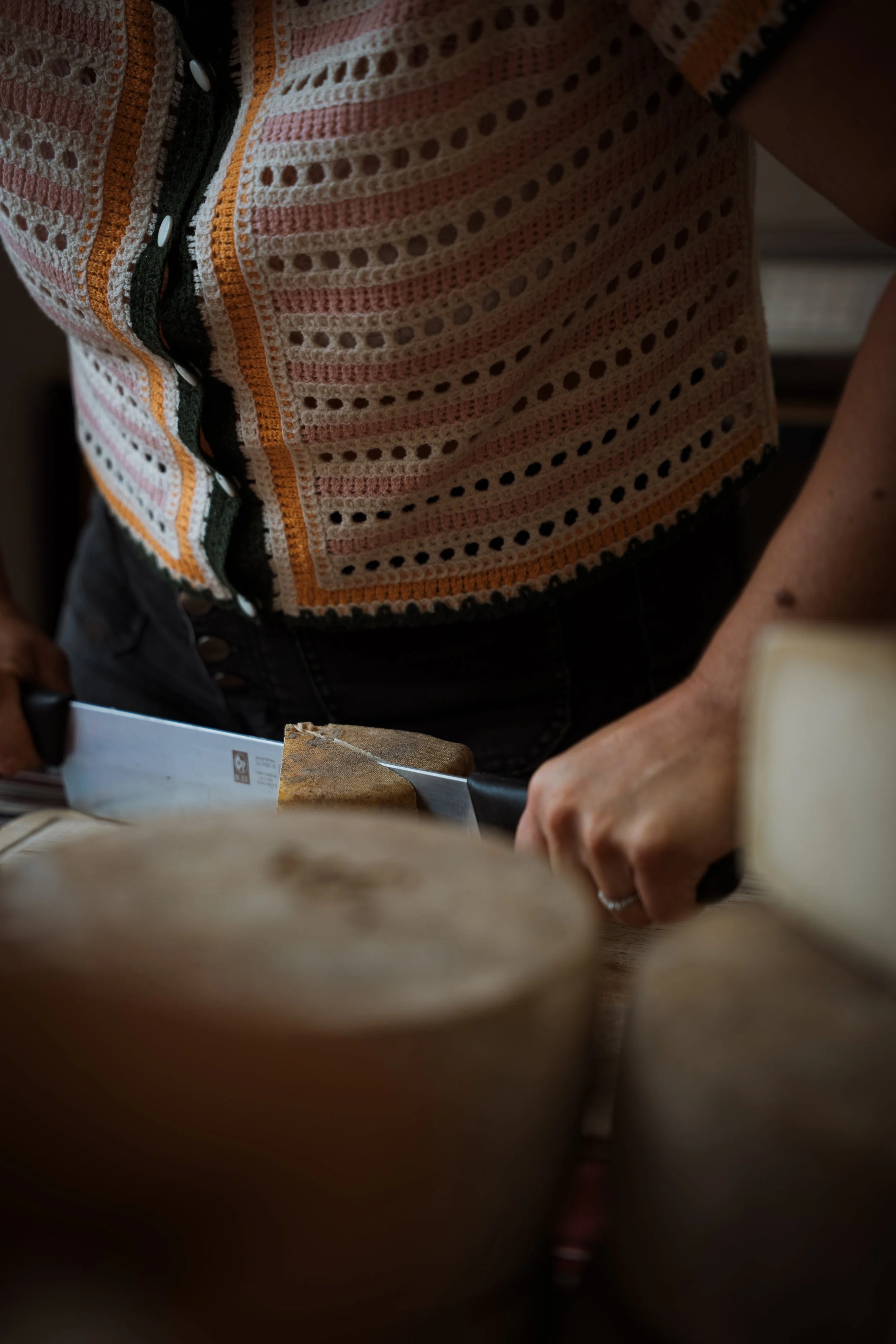 Partie d'une femme portant un vêtement tricoté coloré avec des motifs circulaires, elle tient un couteau de cuisine dans sa main, et travaille sur un plan de travail en bois, probablement en train de cuisiner. La photo est prise de près, avec un focus sur la main et le vêtement.