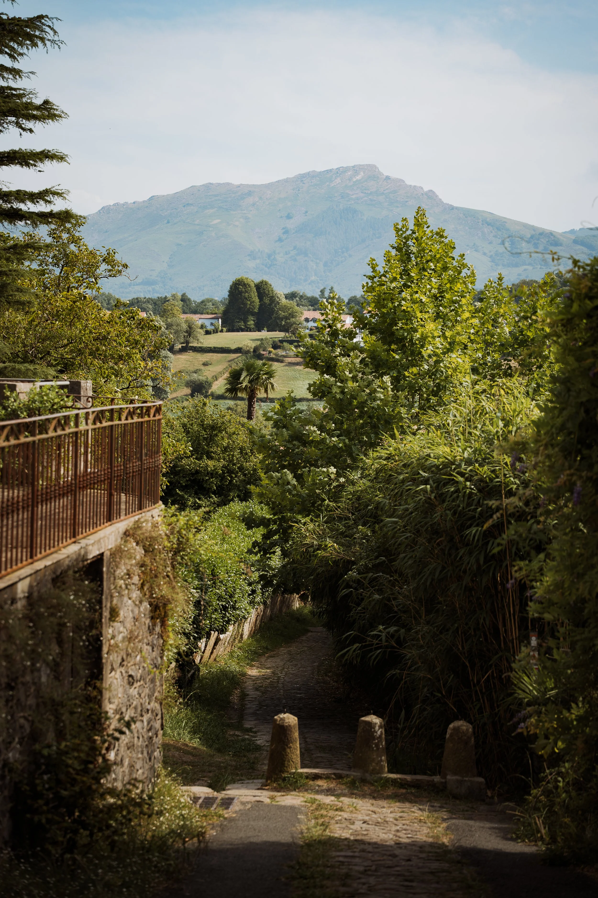 Une ruelle pavée entourée de végétation, avec un mont en arrière-plan sous un ciel clair et partiellement nuageux.