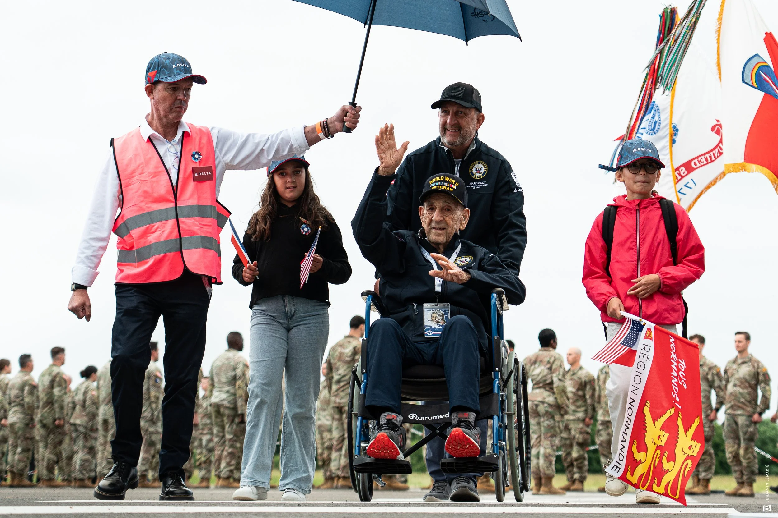 Groupe de personnes, dont un homme en fauteuil roulant, saluant lors d'une cérémonie militaire ou commémorative, avec des soldats en uniforme en arrière-plan.