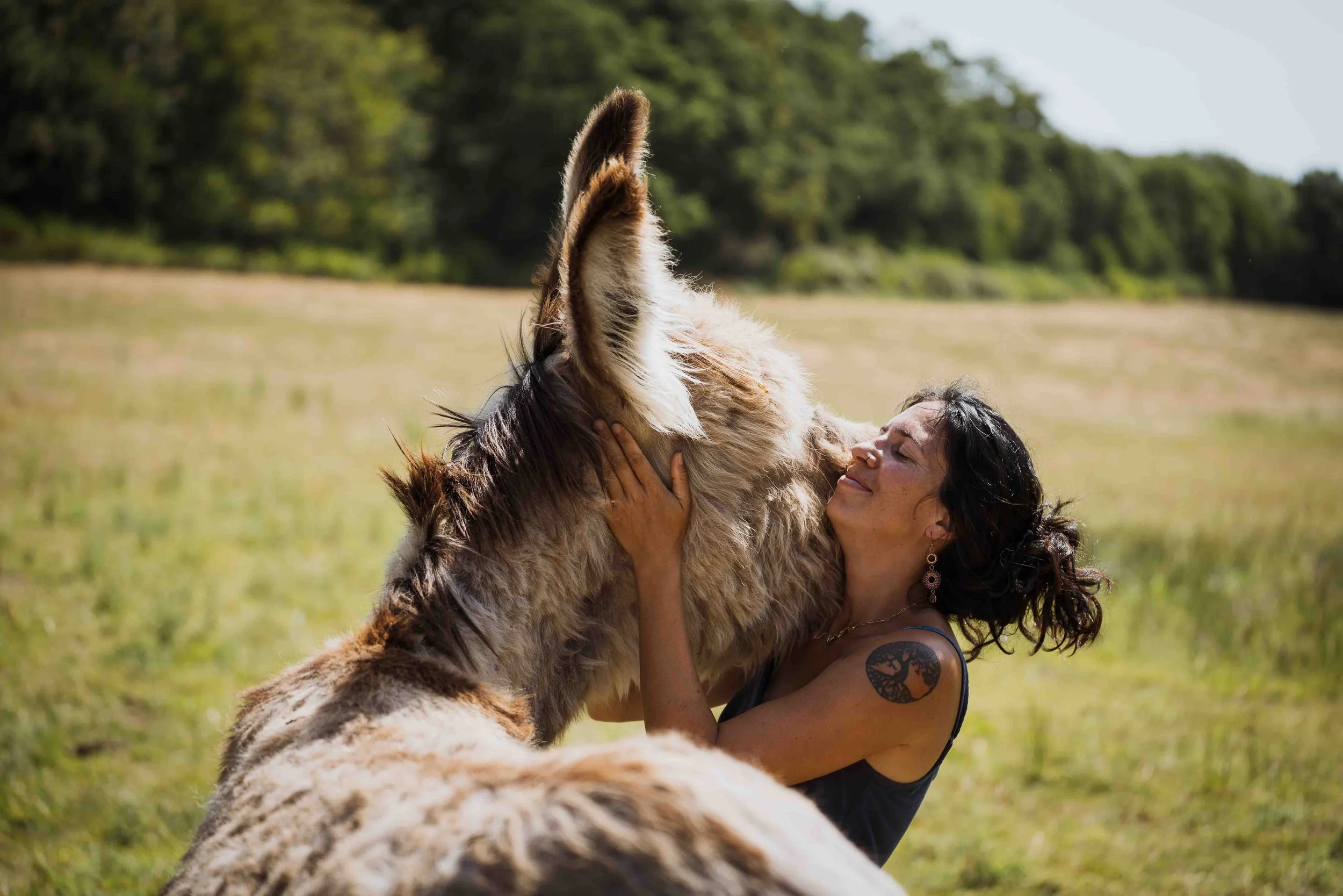 Une femme souriante tient un grand chien de race berger australien dans un champ, entourée d'arbres en arrière-plan.