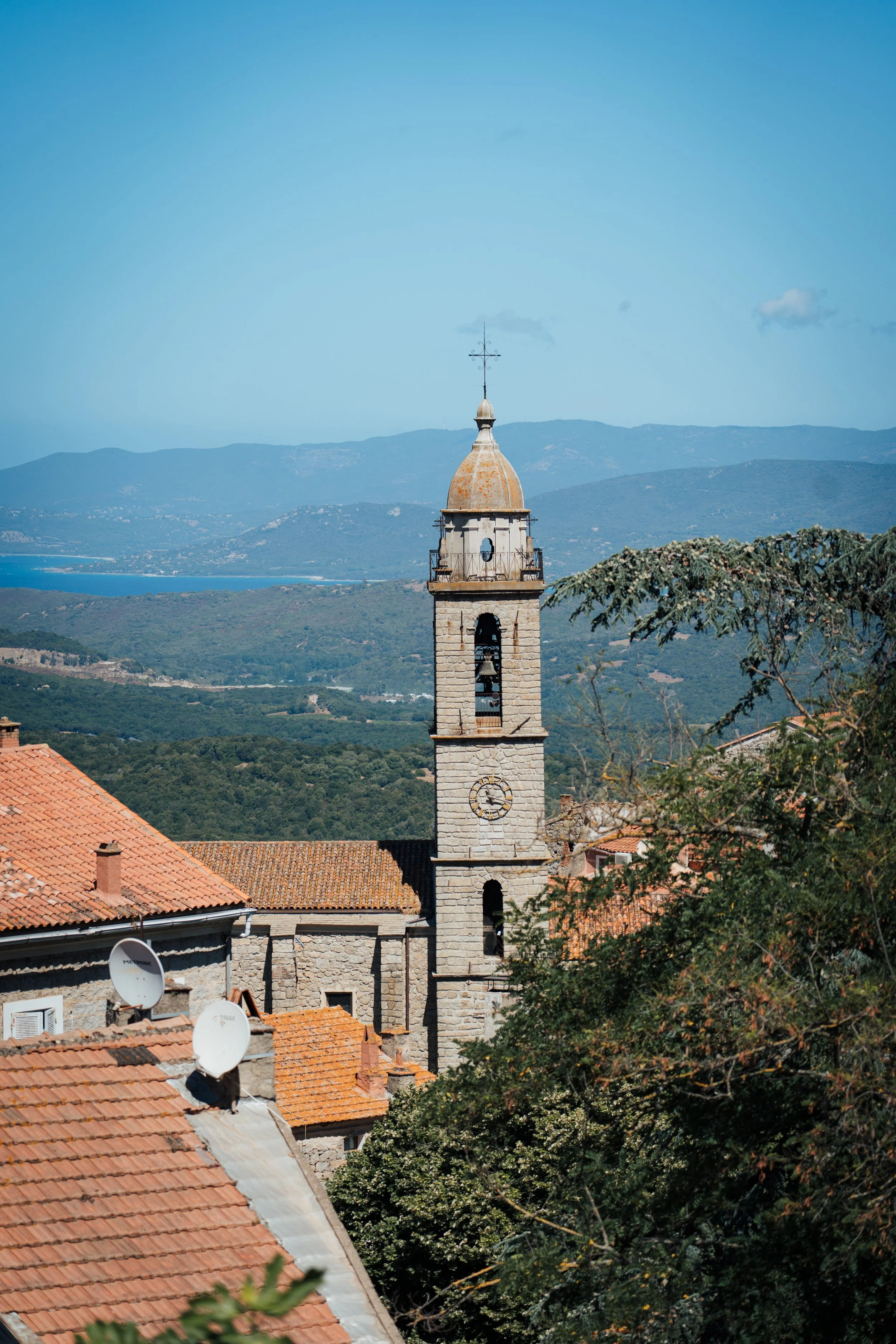 Une église avec un clocher en pierre et un cadran solaire, des toits en tuiles rouges, un paysage de montagnes en arrière-plan et des arbres devant.