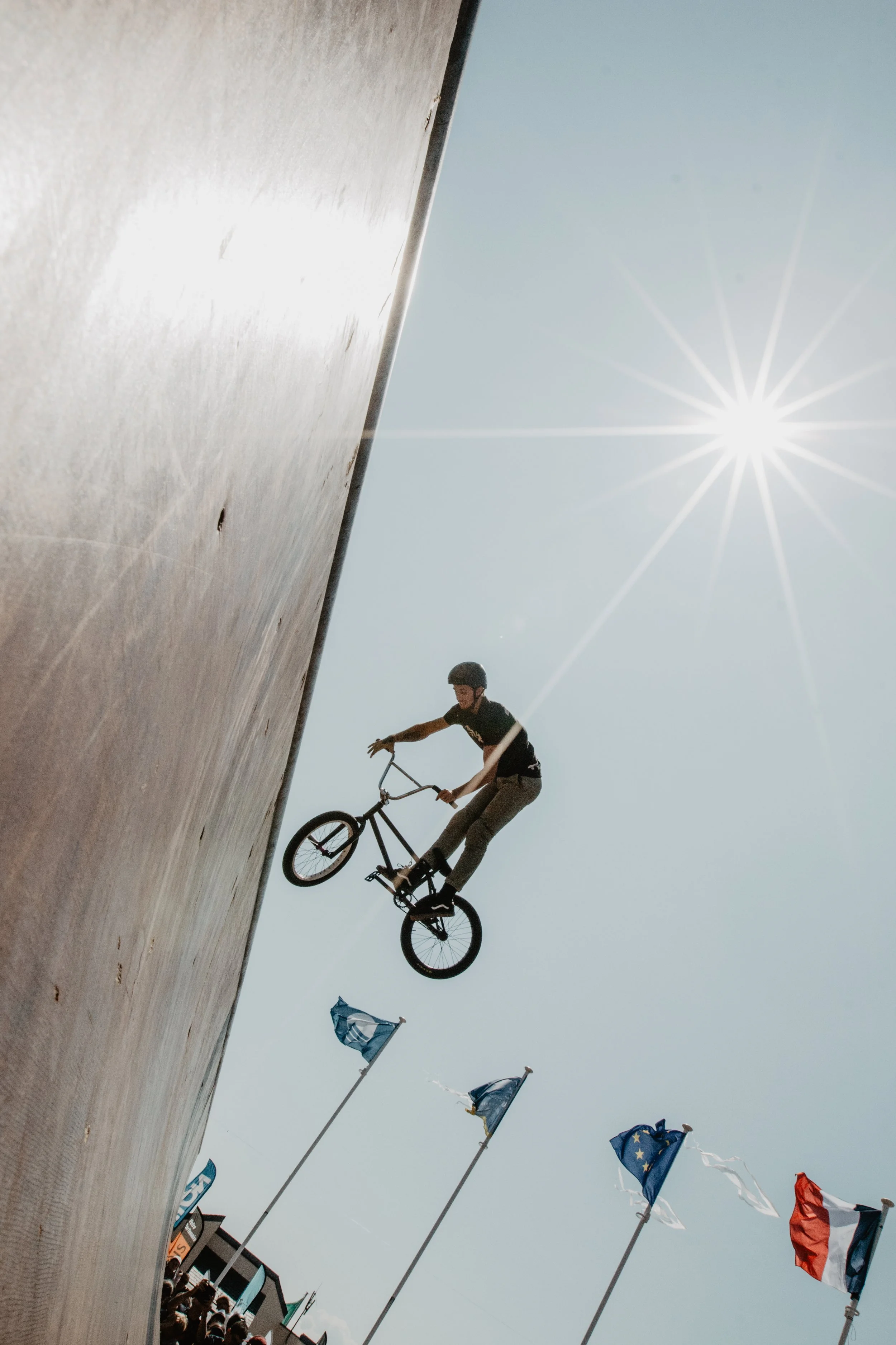 Un cycliste effectue une figure acrobatique avec son vélo en l'air lors d'un spectacle, avec un mur en béton sur la gauche et des drapeaux européens flottants en arrière-plan, sous un ciel ensoleillé.
