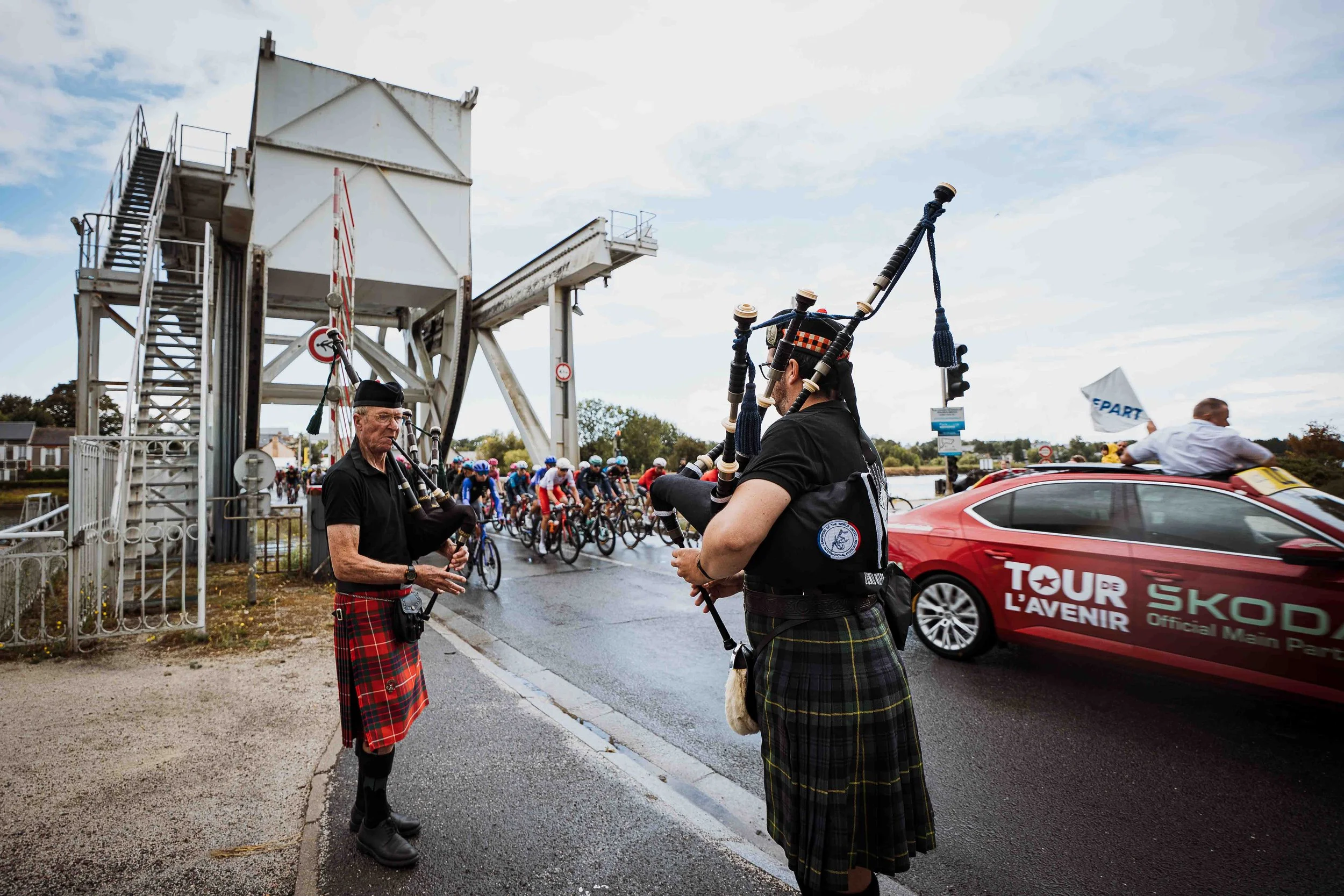 Deux hommes en kilt écossais jouent de la cornemuse devant un groupe de cyclistes lors d'une course ou d'un événement sportif. Un panneau indique 'Tour de l'avenir'.