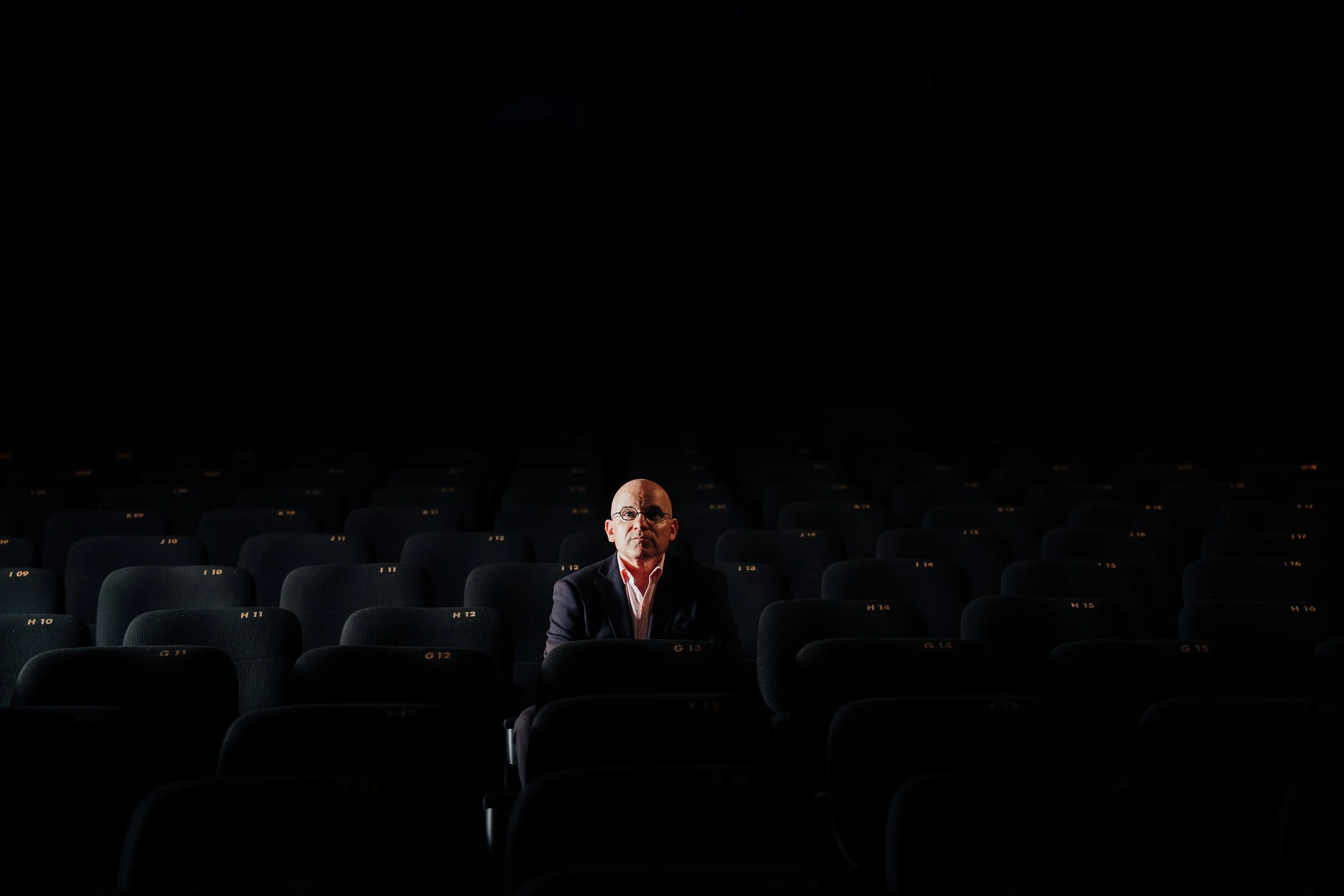 Un homme seul assis dans une salle de cinéma sombre avec des sièges numérotés, portant des lunettes et une chemise blanche, avec un look sérieux.