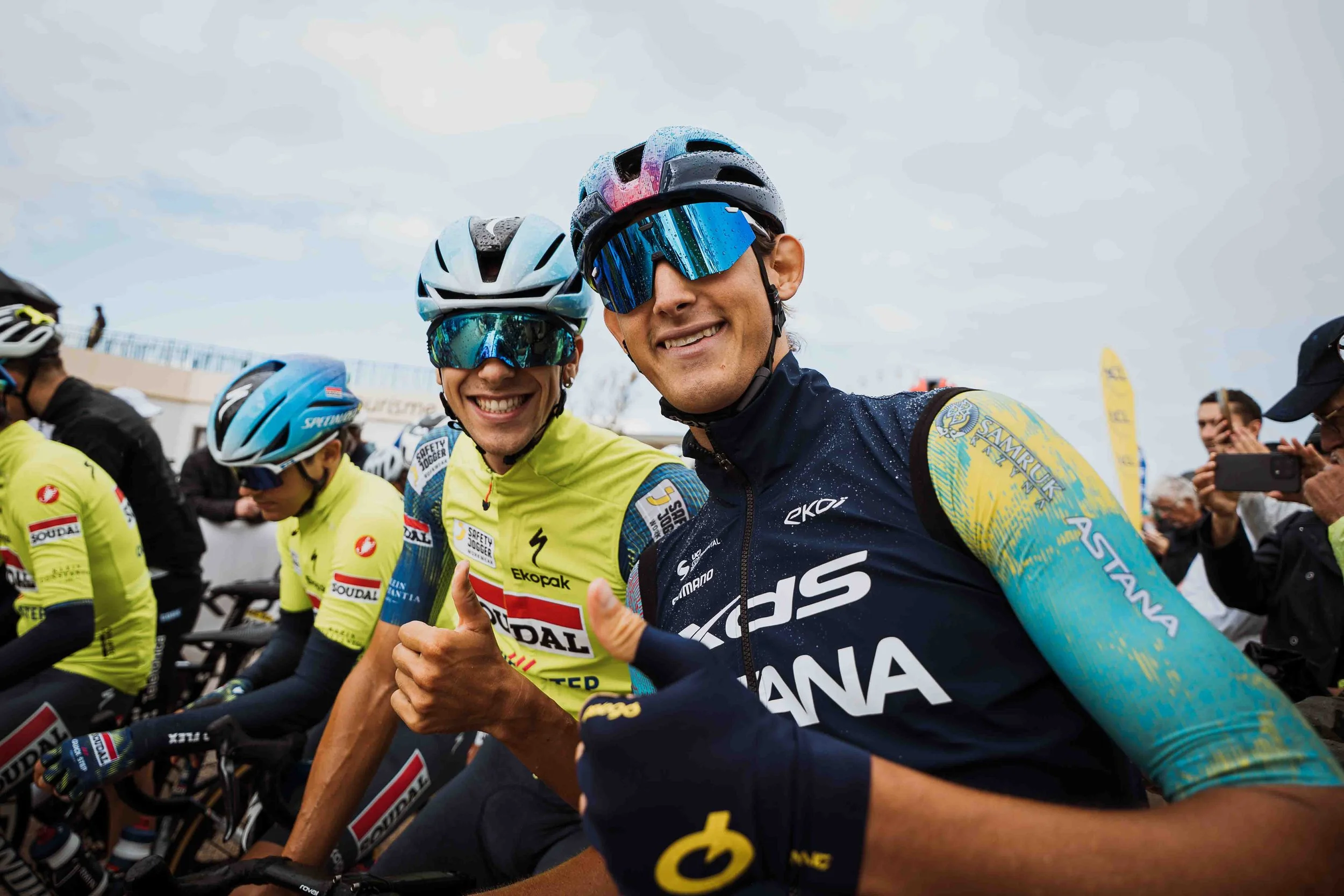 Des cyclistes en uniforme jaune et noir avec casques, souriant et faisant un geste de voeux, lors d'une course ou d'un événement sportif, avec des spectateurs en arrière-plan.