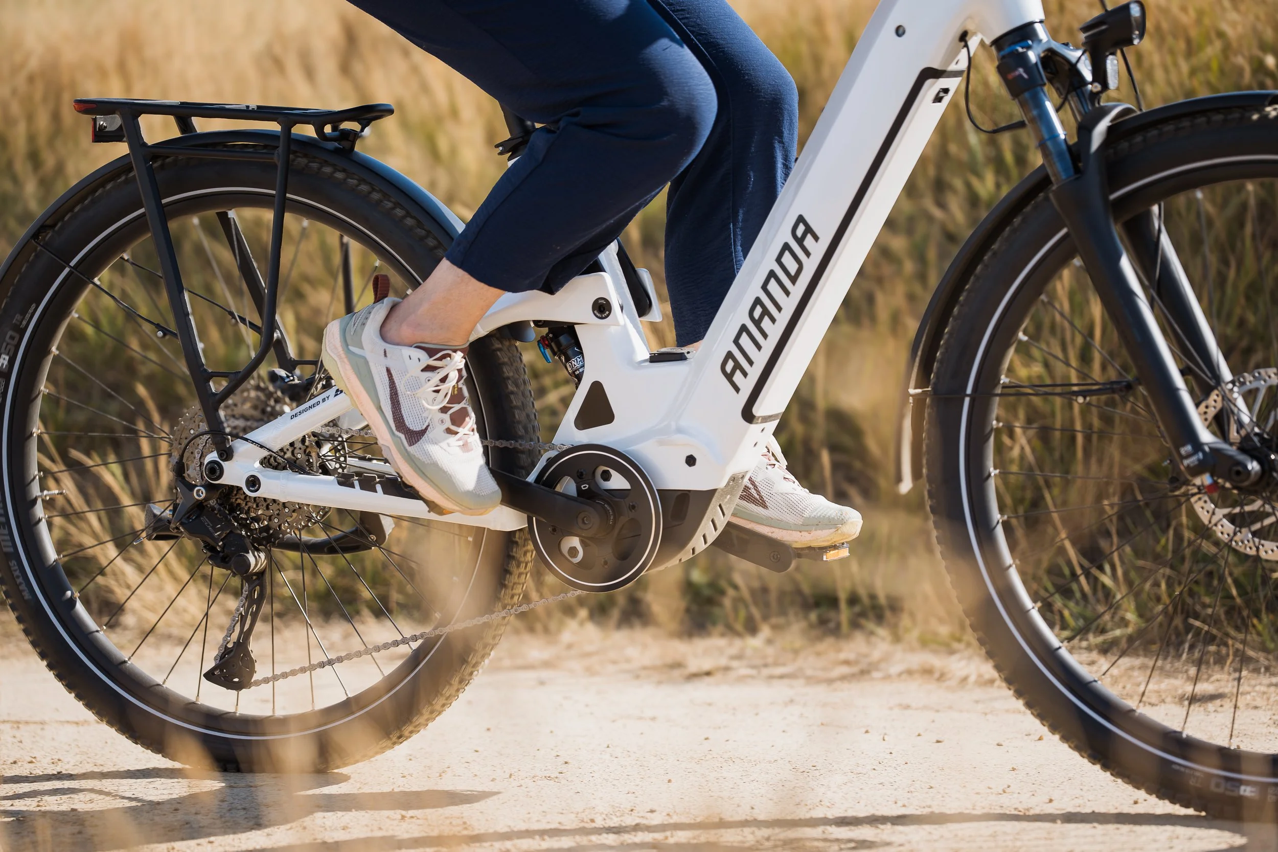 Vue rapprochée d'un vélo électrique blanc avec le nom 'Aventon' sur le cadre, qui est en mouvement sur un chemin de terre avec un arrière-plan naturel de végétation sèche.
