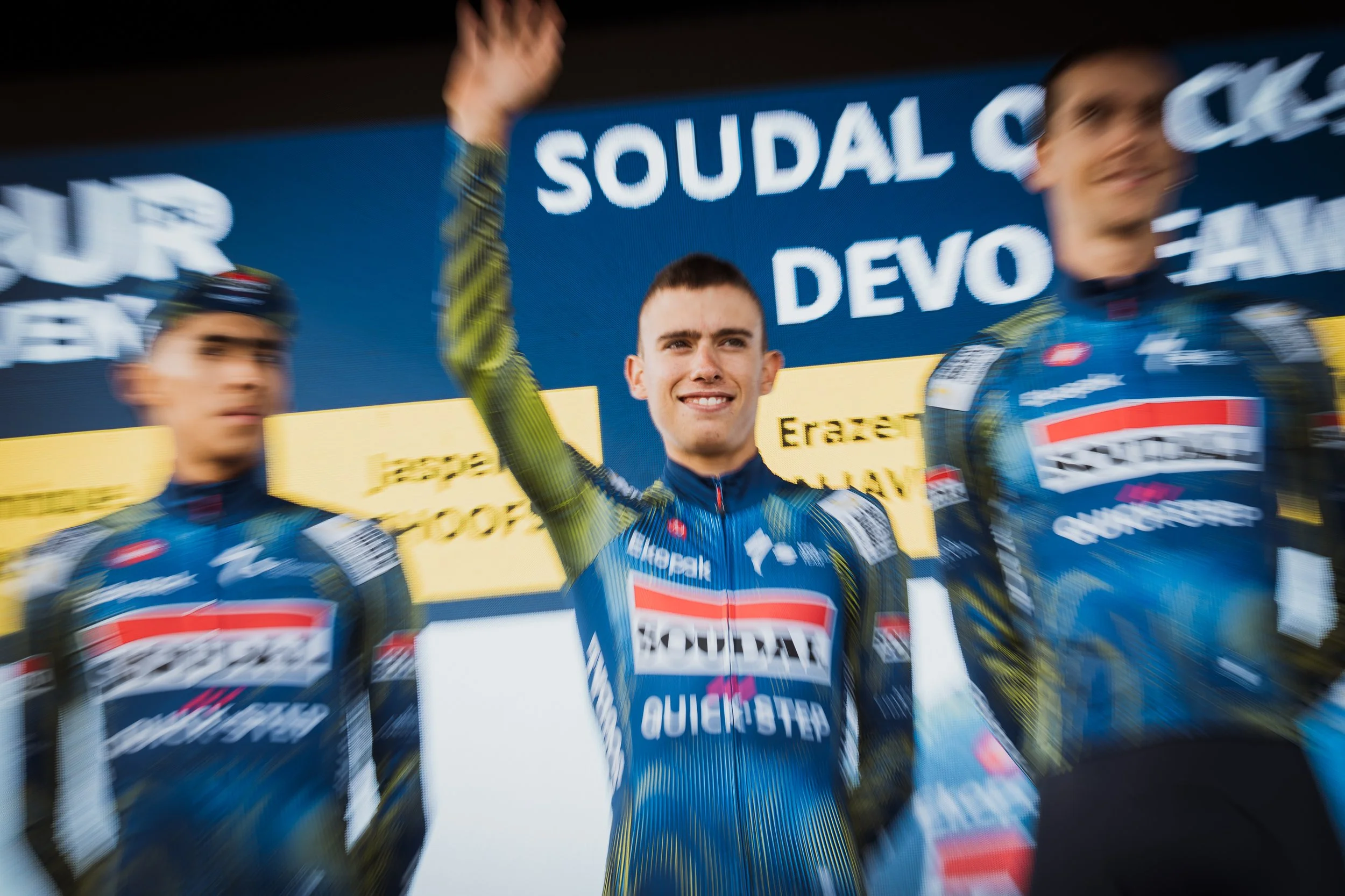 Trois cyclistes en tenue de course sur le podium, la personne centrale sourit et lève la main, devant un panneau avec le texte "SOUDAL" et des autres inscriptions, la scène semble être une cérémonie de remise de prix.