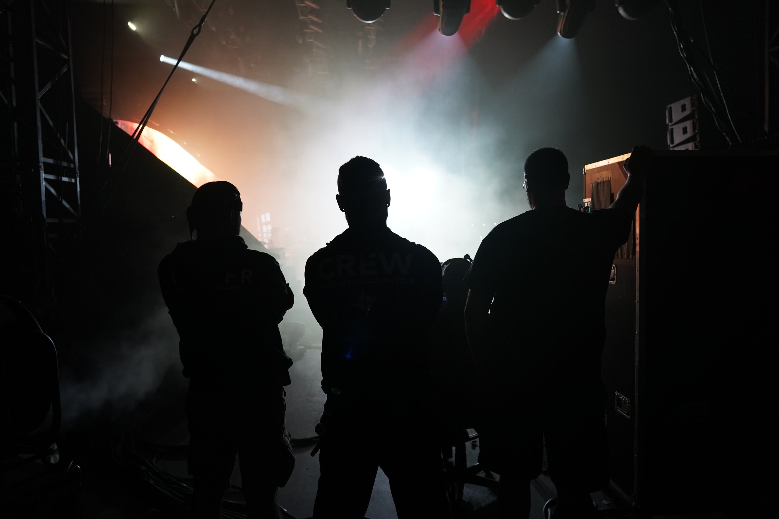 Silhouettes de trois personnes dans une salle sombre avec un éclairage brillant à l'arrière, suggérant une scène de préparation pour un spectacle ou un événement.
