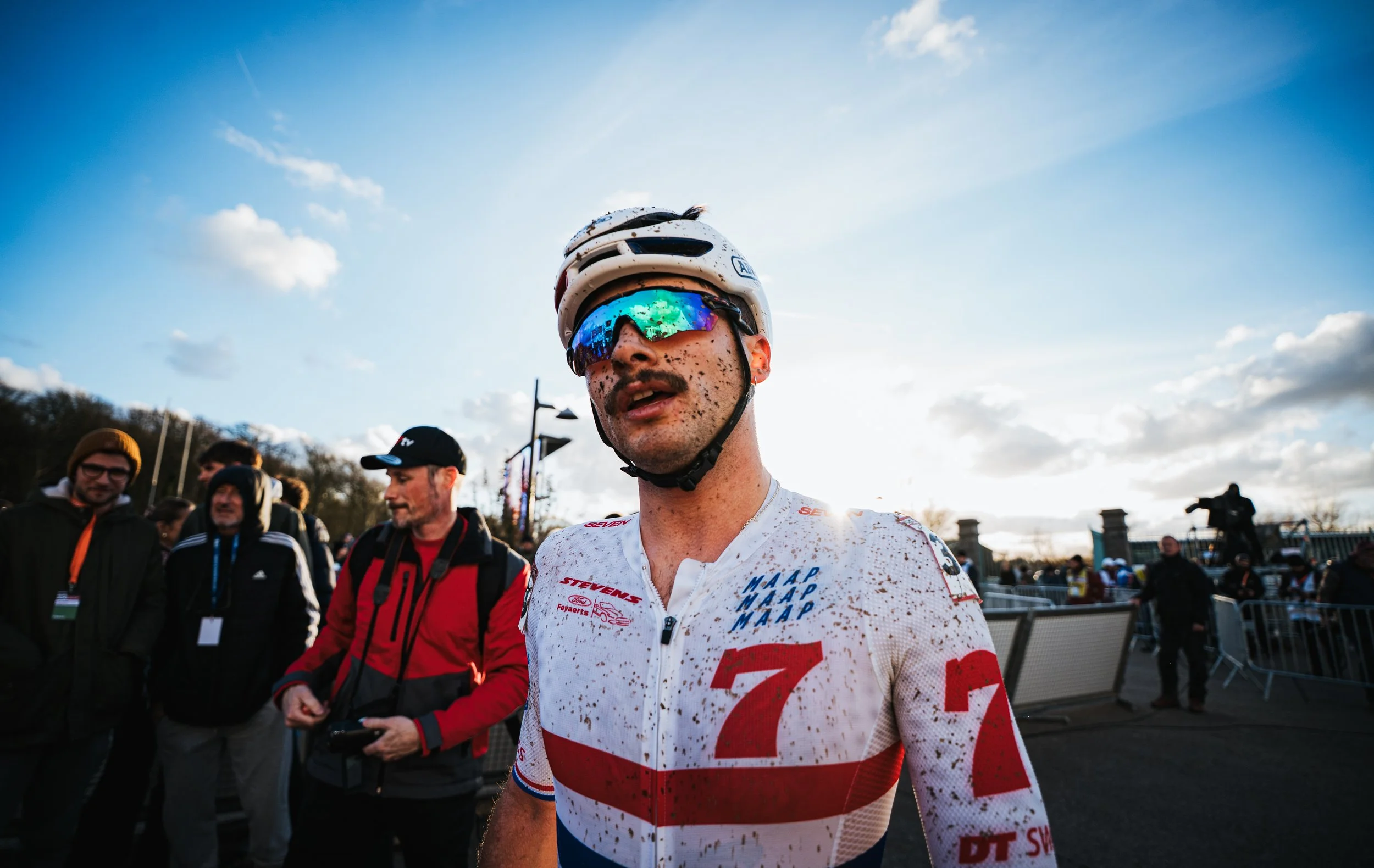 Un cycliste portant un casque blanc et des lunettes réfléchissantes se tient parmi un groupe de personnes, avec un ciel nuageux en arrière-plan. Le cycliste est couvert de boue et porte un maillot de cyclisme blanc avec des inscriptions rouges et bleues.